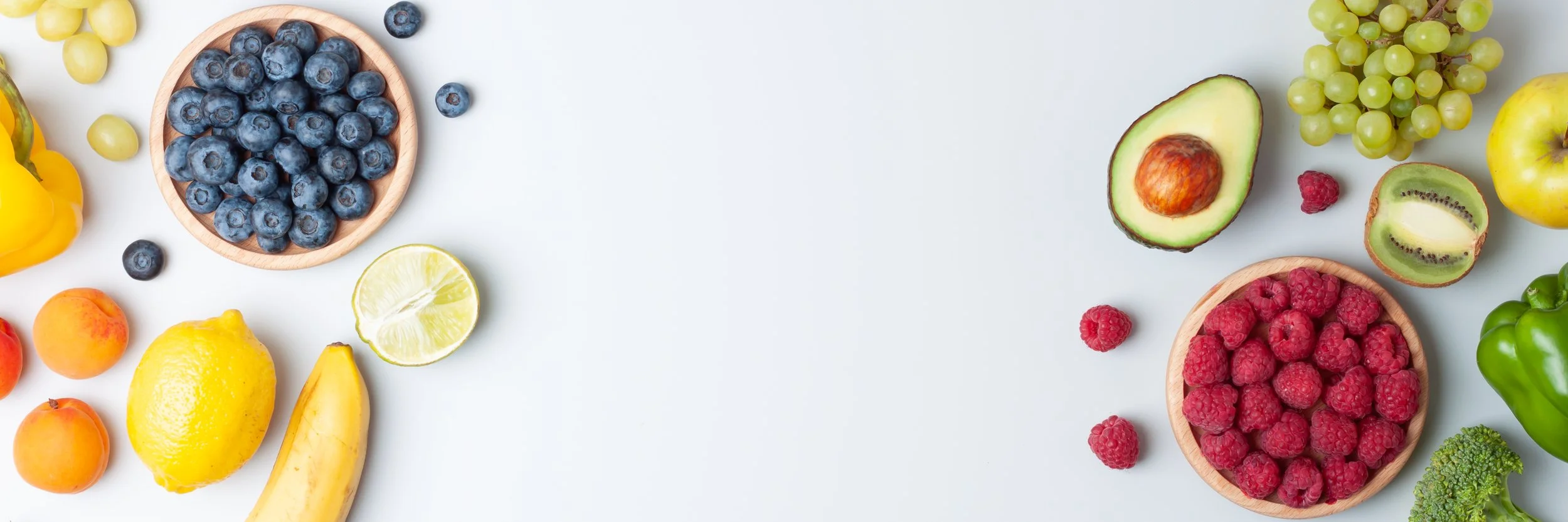 Assorted fresh fruits including blueberries, raspberries, avocado, green grapes, yellow lemon, banana, yellow bell pepper, peach, kiwi, broccoli, and lime, arranged on a white background