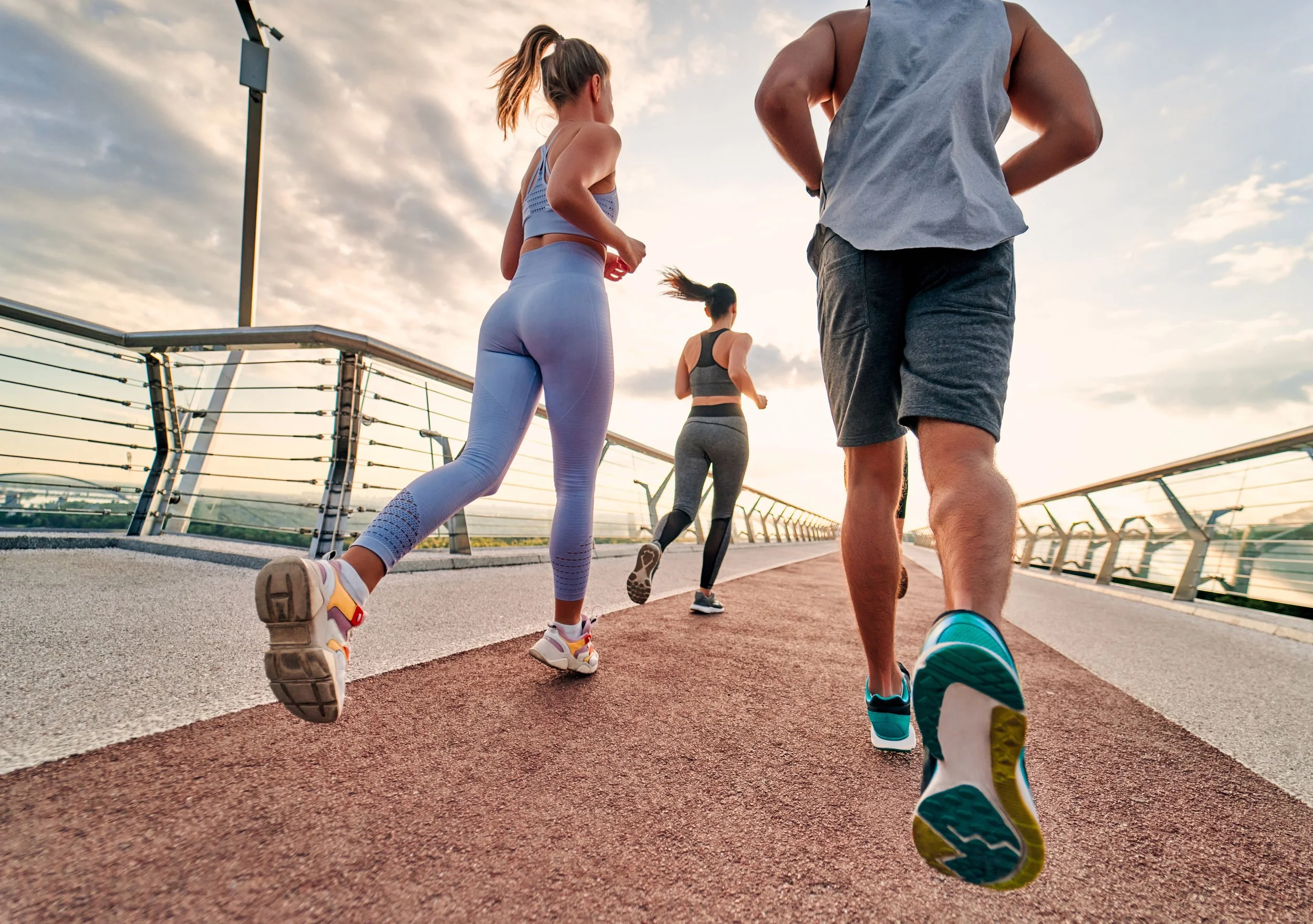 Four people running on a bridge at sunset, viewed from behind. Two women and two men are running, dressed in athletic wear. The sky is partly cloudy with a warm glow from the setting sun.