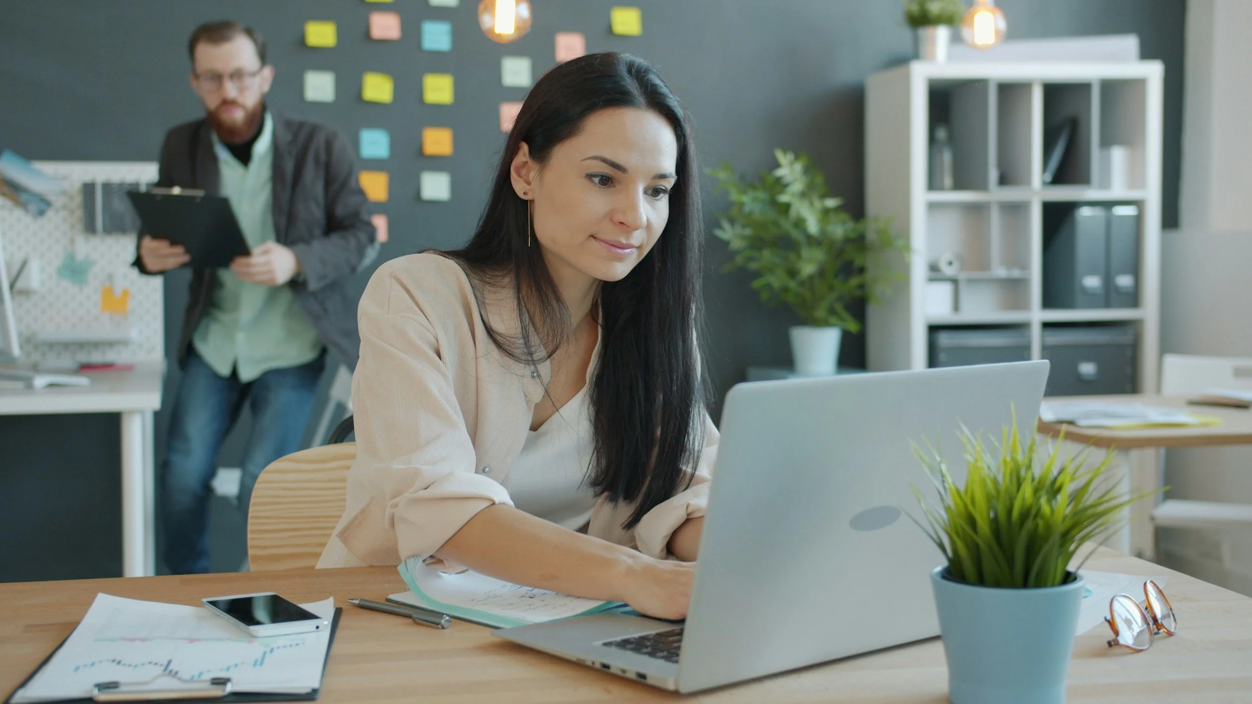 A woman working on a laptop at a desk with a plant, glasses, and a phone nearby; in the background a man holding a clipboard in an office.