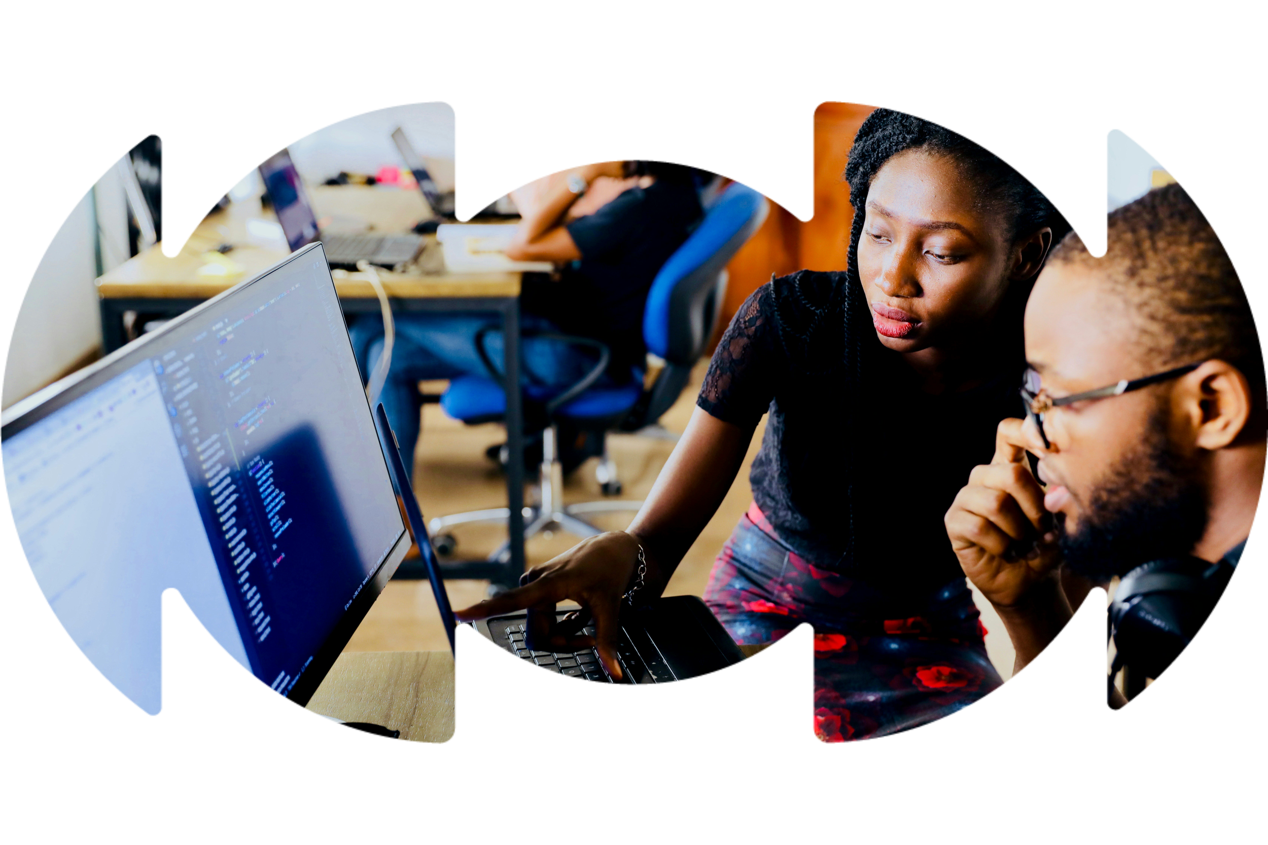Two women working together on a computer in an office environment, with other employees working at desks in the background.