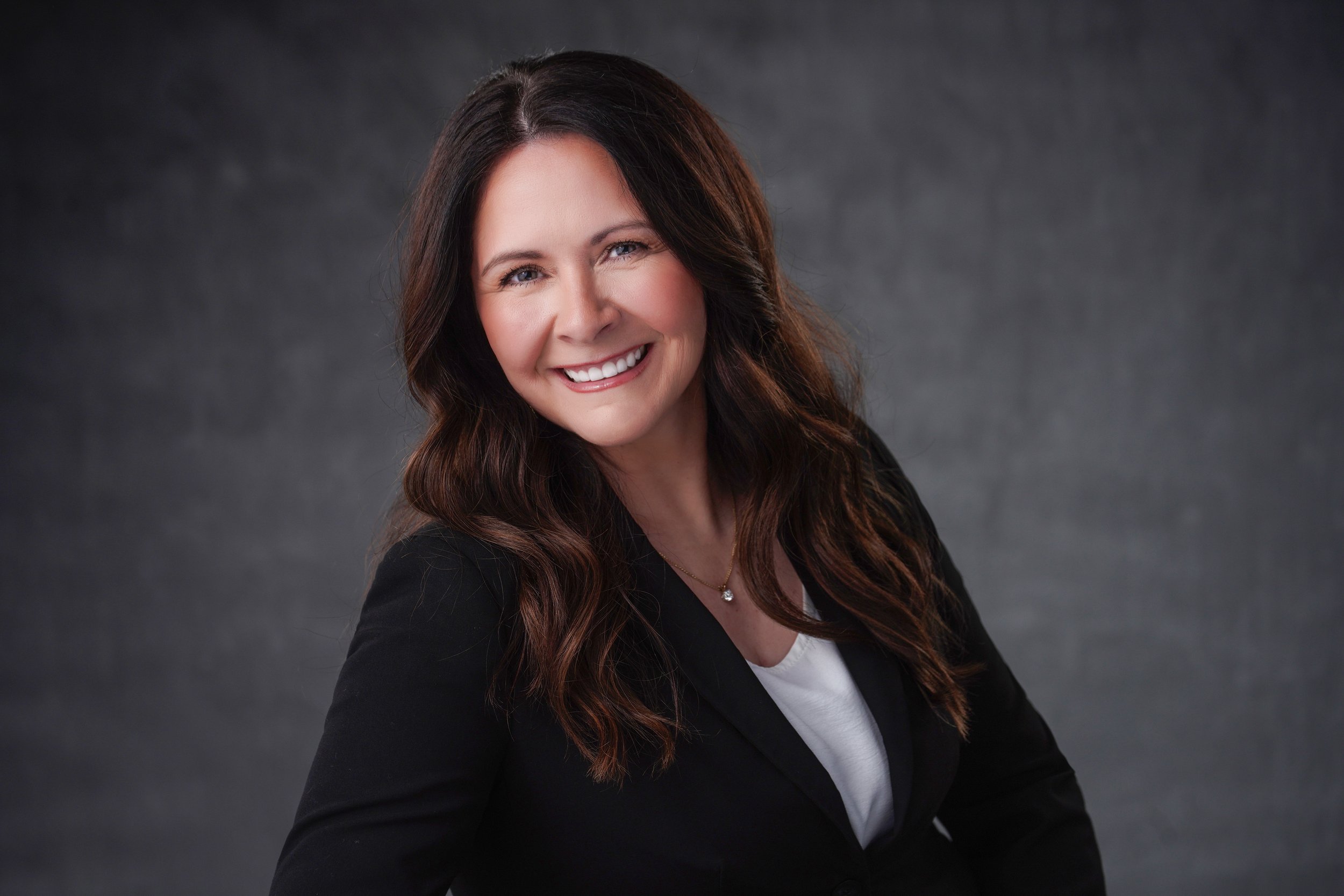 Headshot of a woman with long wavy brown hair, smiling, wearing a black blazer over a white top, against a dark gray background.