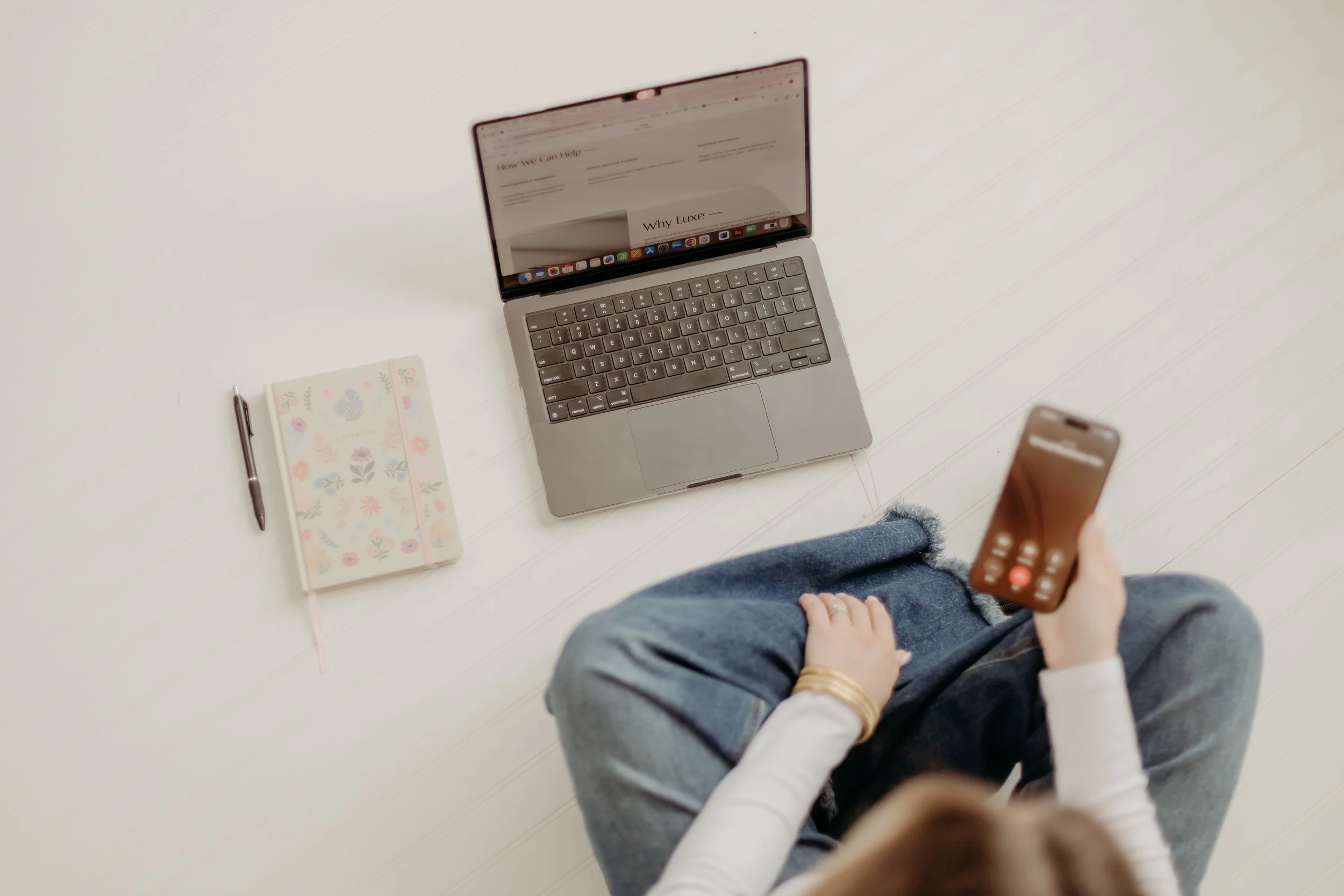 A person sitting on a white floor with a MacBook laptop, a floral notebook, a pen, and holding a smartphone.