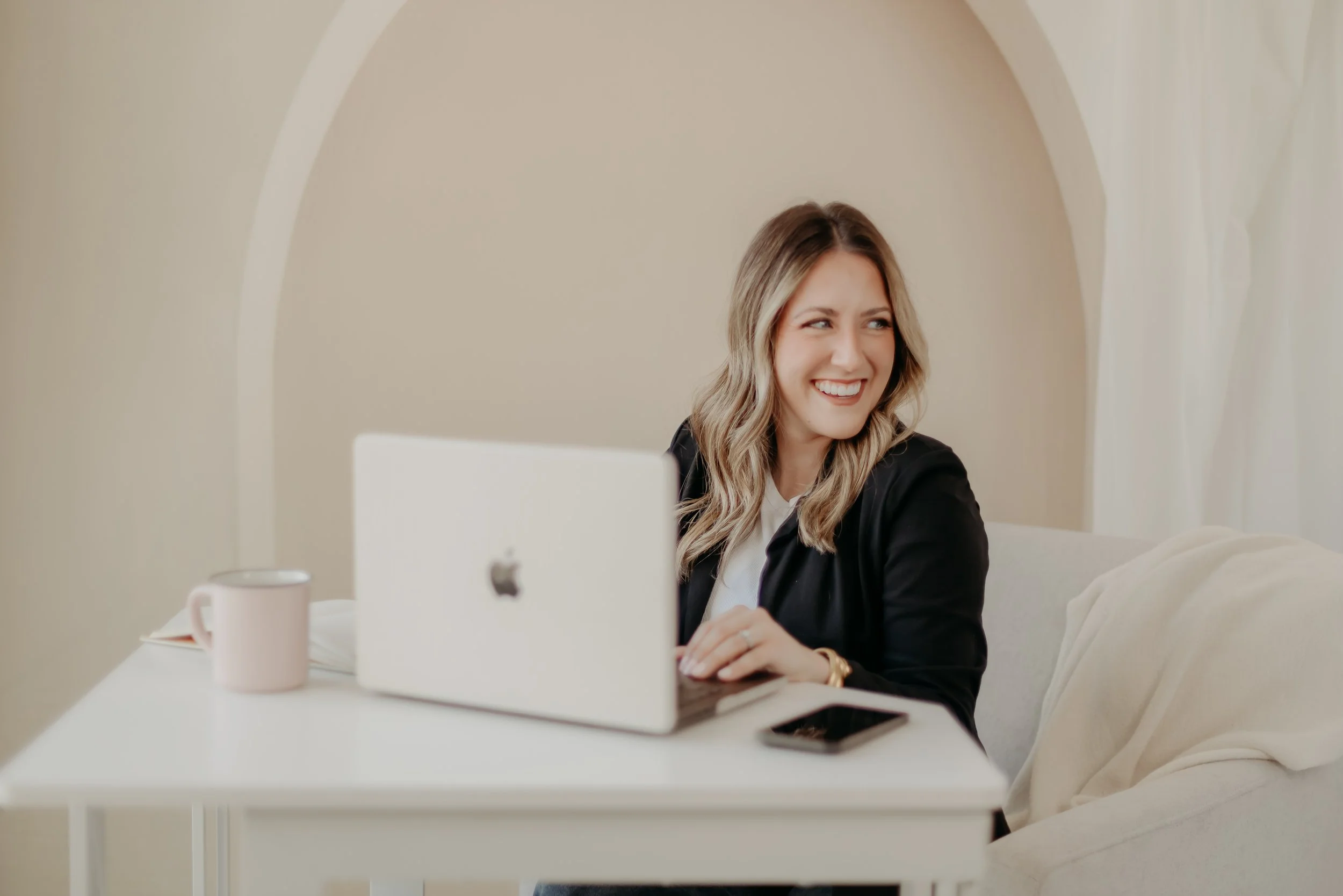 A woman sitting at a white desk with a MacBook, a smartphone, a pink coffee mug, and a notebook, smiling and looking to her right in a bright room.