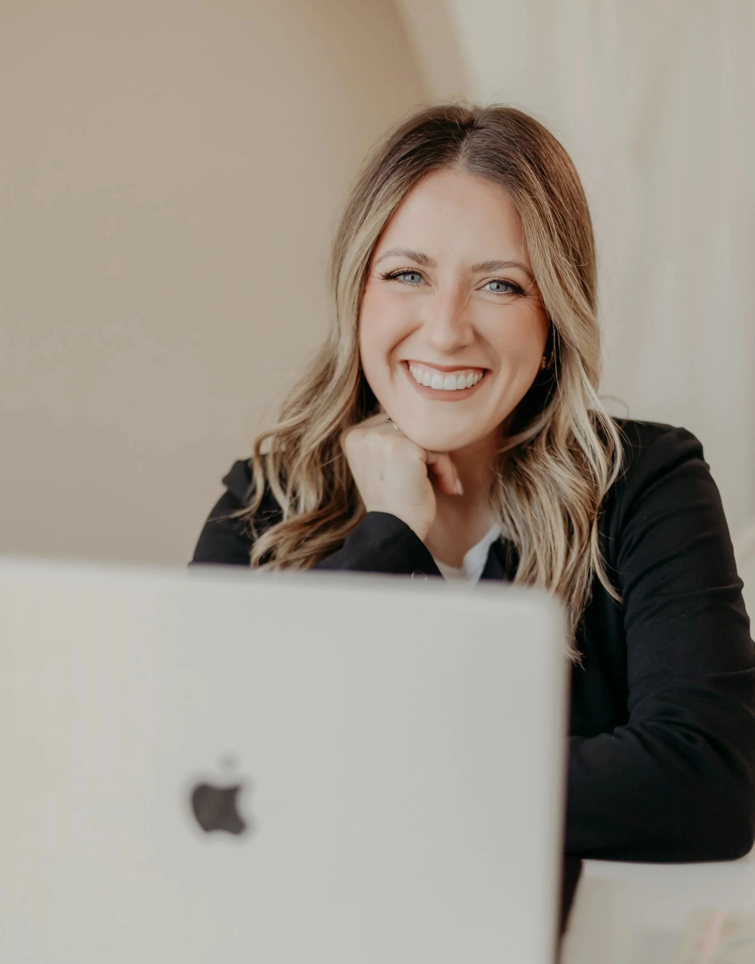 A smiling woman with light brown, wavy hair and blue eyes, sitting at a desk with an iMac computer, resting her chin on her hand, in an indoor setting.