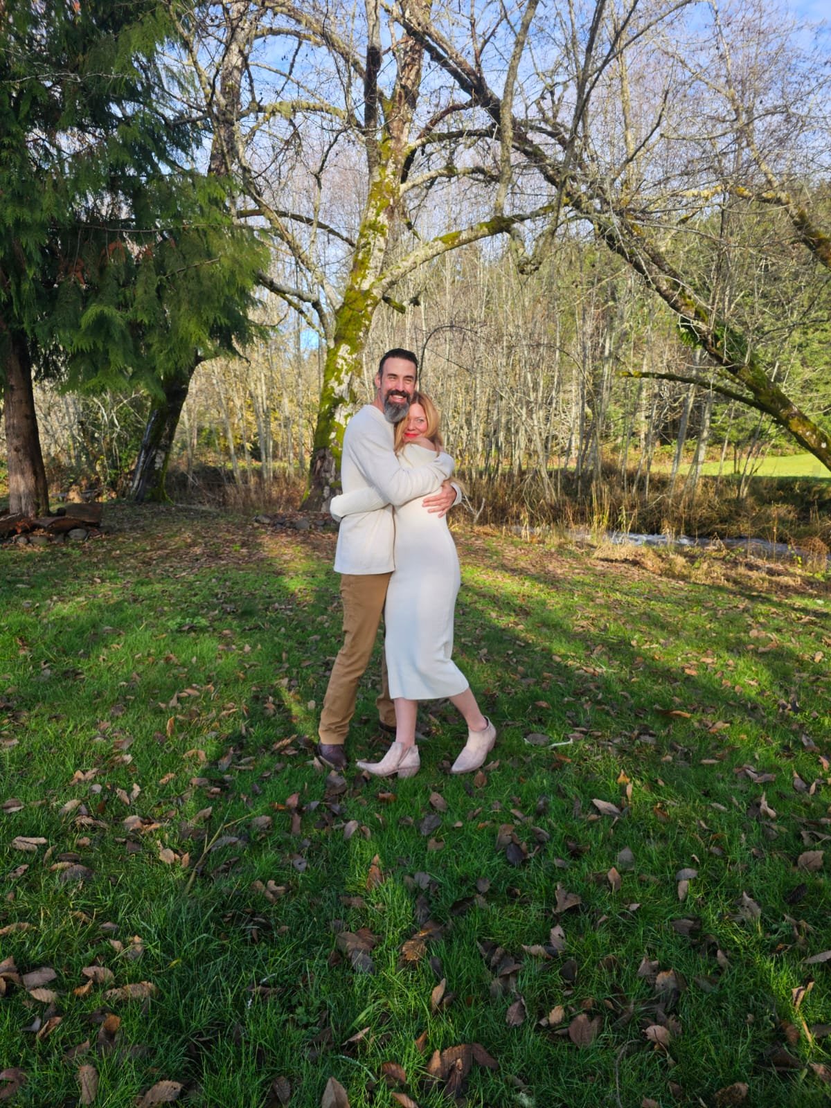 A man and woman hugging outdoors in a grassy area with trees, some leaves on the ground, and a small creek visible in the background.