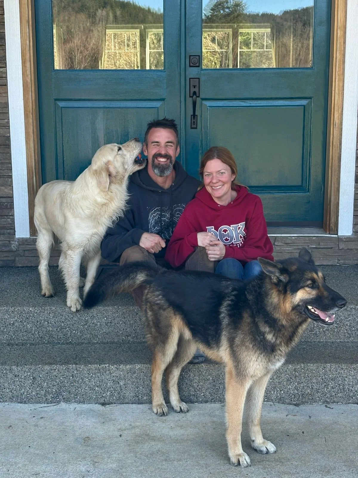 A man and woman sitting on the porch of a house with two dogs, one Golden Retriever and one German Shepherd, in front of a large green double door with windows reflecting trees and sky.