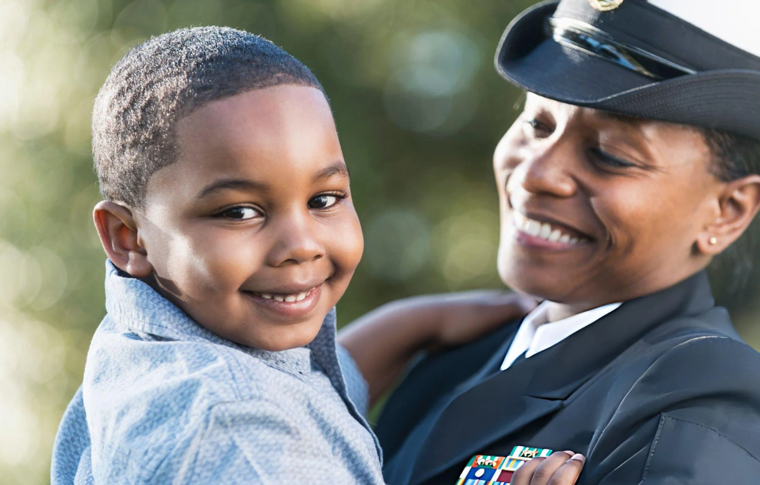 A smiling young boy is being held in the arms of a woman in a military uniform with medals, both smiling and outdoors with blurred greenery in the background.