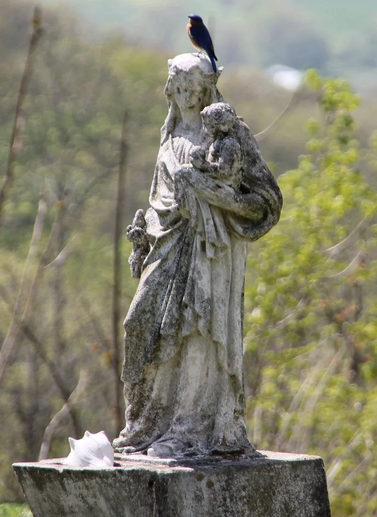 A weathered stone statue of a woman holding a child, with a small bird perched on the top of her head and a shell on the base in an outdoor setting with greenery.
