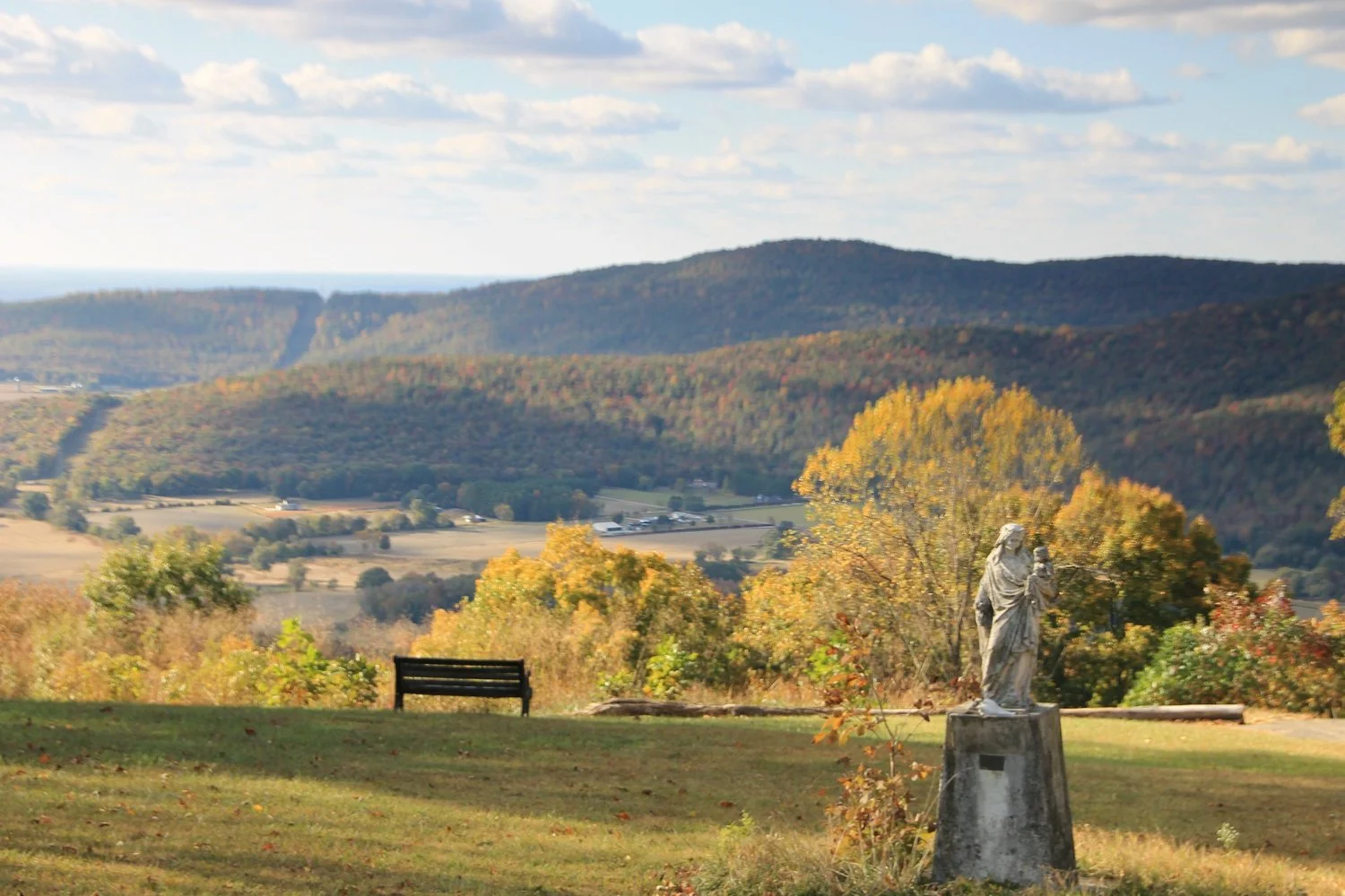 A scenic landscape with rolling hills in the background, colorful autumn trees, a statue of a religious figure holding a child, and a park bench in the foreground.