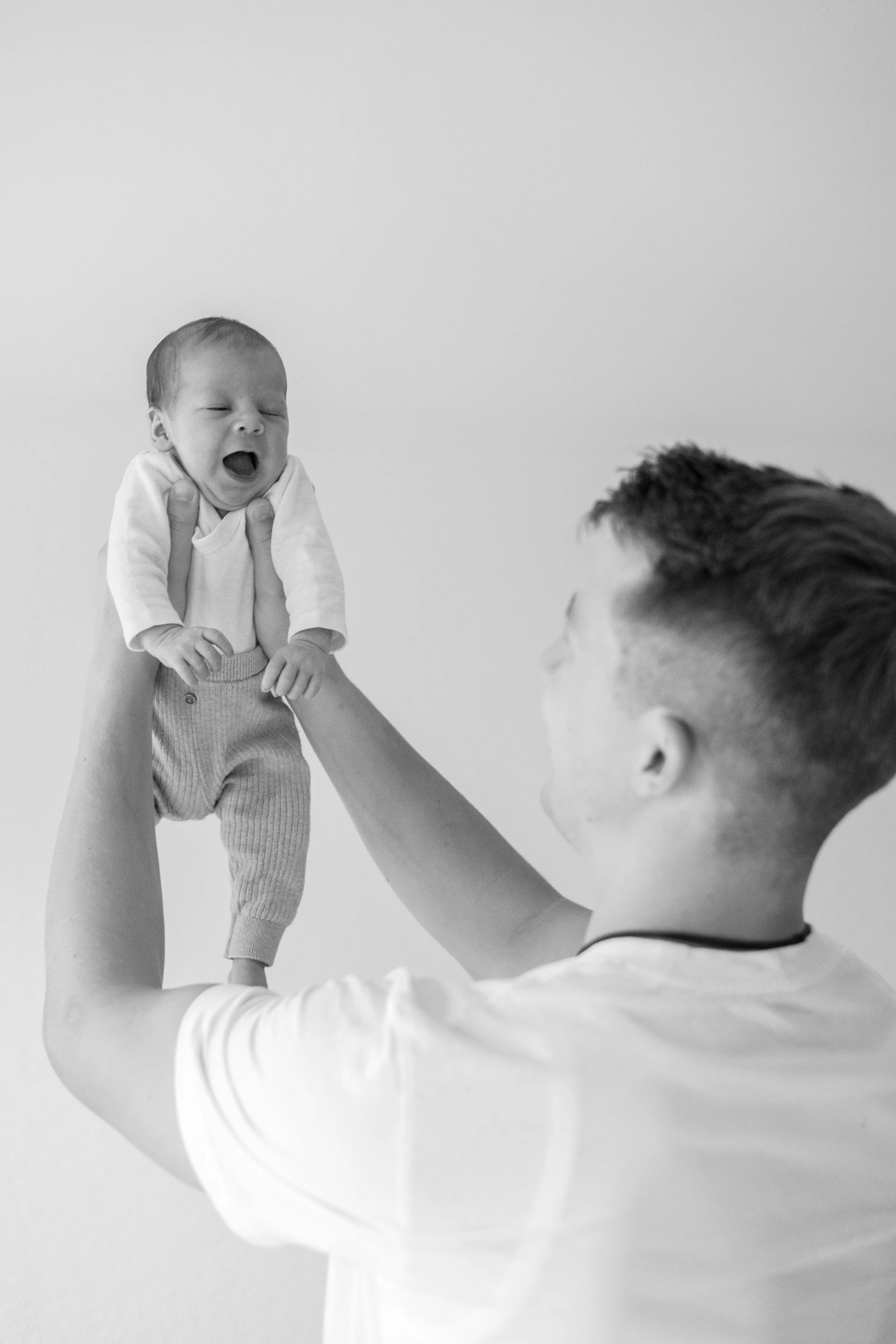 Ein Mann hält ein schreiendes Baby in die Luft, die Szene ist in Schwarz-Weiß. Der Mann trägt ein weißes T-Shirt, das Baby trägt eine weiße Kleidung und graue Strümpfe.