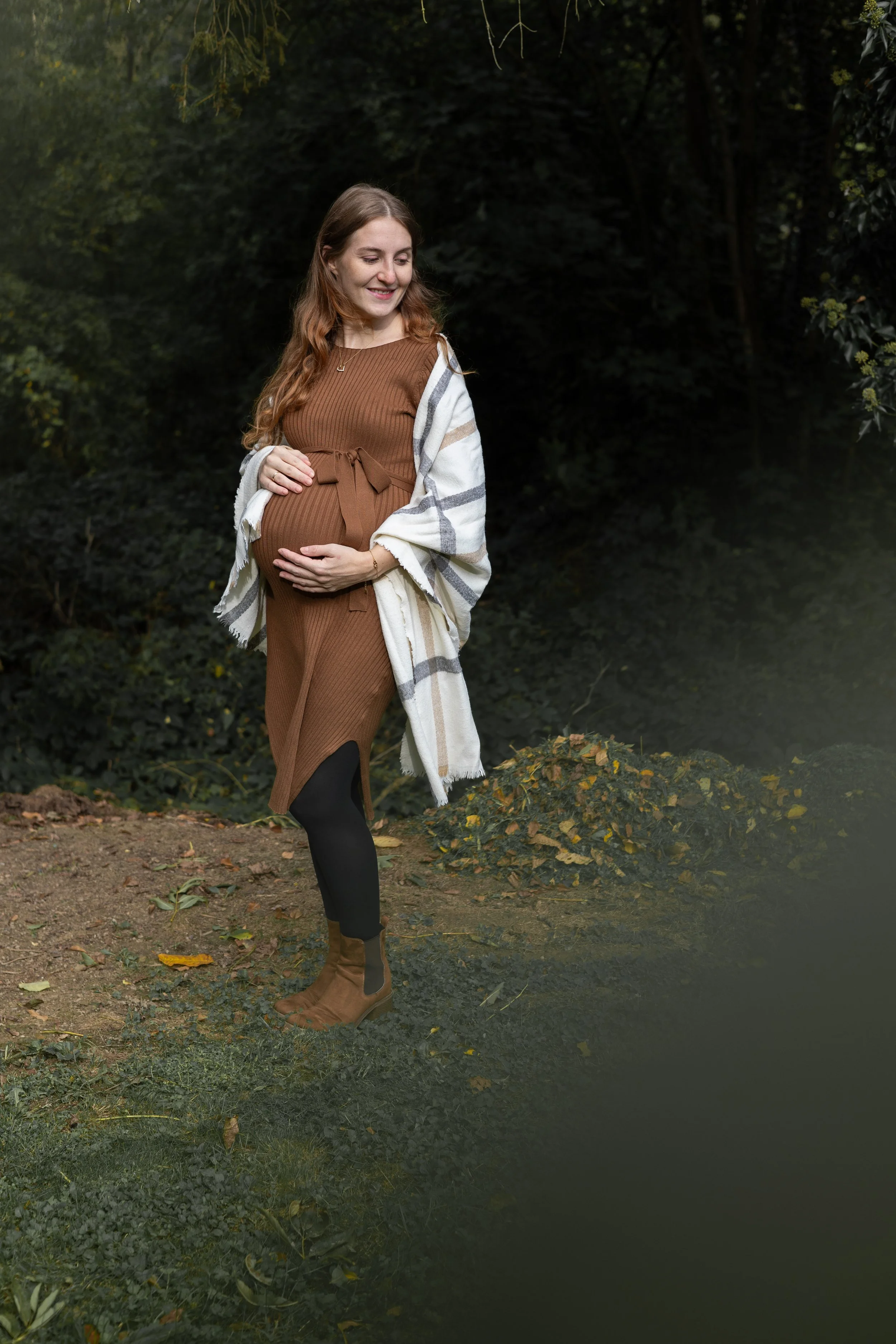 Eine schwangere Frau mit langen braunen Haaren, gekleidet in einem braunen Kleid und braunen Stiefeln, steht in einem dunklen Waldumfeld und hält ihren Babybauch mit beiden Händen. Sie lächelt und schaut nach unten.