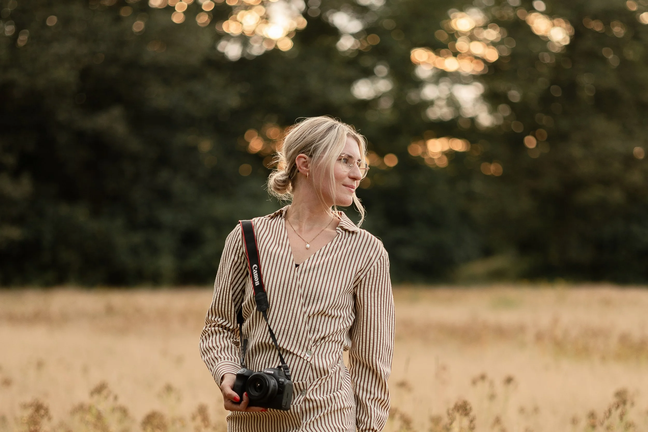 Eine junge Frau mit Brille, blonden Haaren in einem Dutt, trägt eine gestreifte Bluse mit Kragen, hält eine Kamera in der Hand und steht in einer offenen, von Bäumen umgebenen Landschaft bei Sonnenuntergang.
