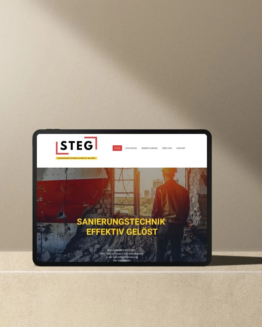 Tablet auf einem Tisch zeigt die Webseite eines Bau- oder Sanierungsunternehmens mit dem Namen STEG und einem Bild eines Bauarbeiters vor einer zerstörten Wand.