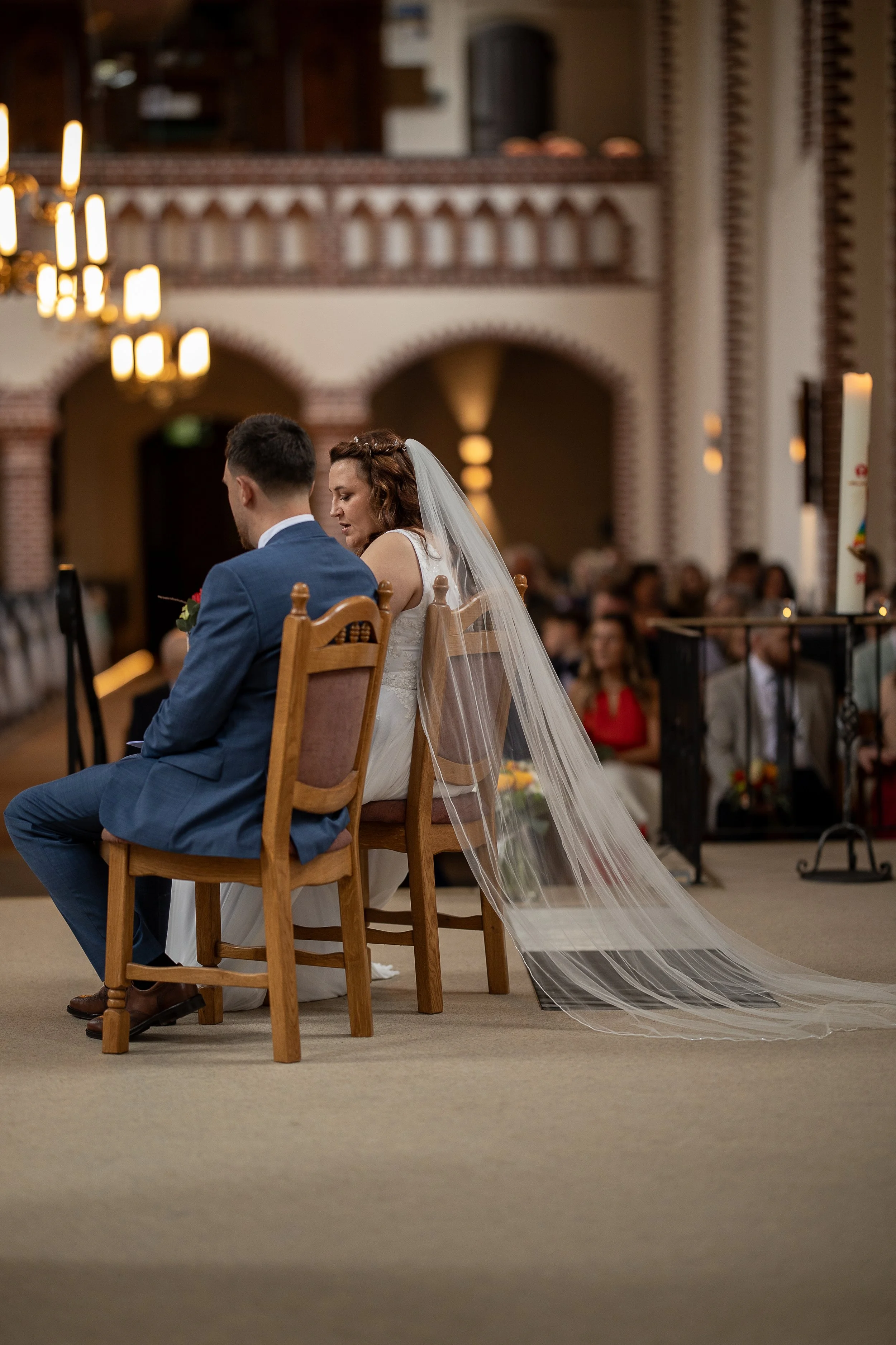 Ein Brautpaar während einer Trauung in einer Kirche, sitzend in Holzstühlen, mit Blick nach vorne, die Braut trägt ein weißes Kleid und einen Schleier, der Bräutigam trägt einen blauen Anzug.