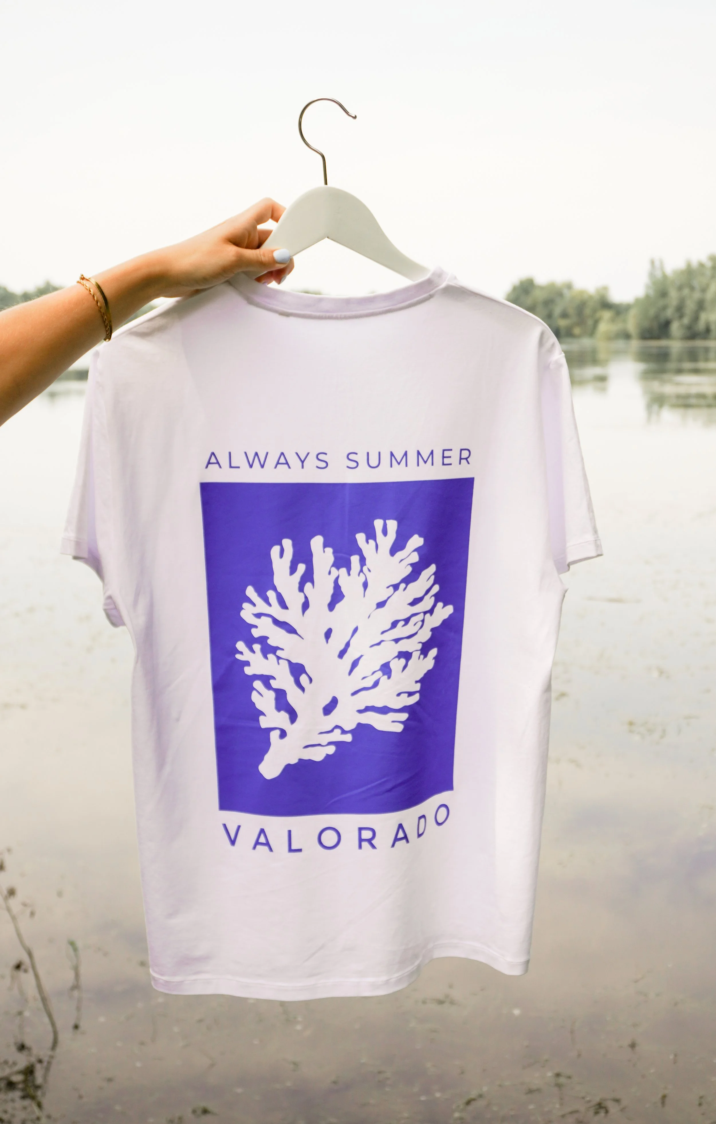 Ein weißes T-Shirt mit einem blauen Design und dem Text „ALWAYS SUMMER“ sowie einer Koralle, es hängt an einem weißen Kleiderbügel, im Hintergrund eine Wasserlandschaft mit Bäumen.