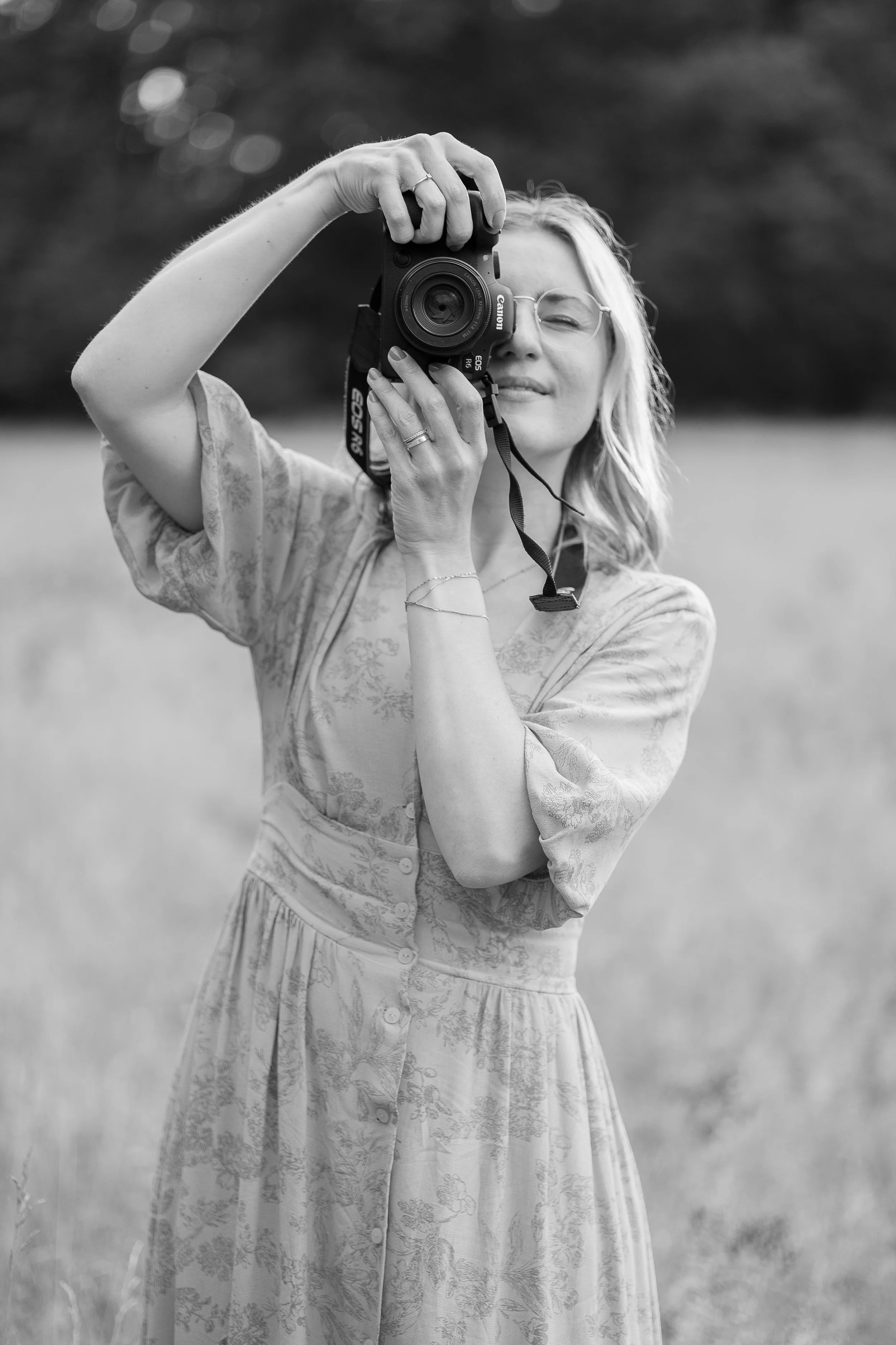 Frau fotografiert mit einer Kamera im Freien. Sie trägt eine leichte, langärmlige Bluse oder Kleid und trägt eine Brille. Der Hintergrund ist unscharf mit Bäumen oder Sträuchern.