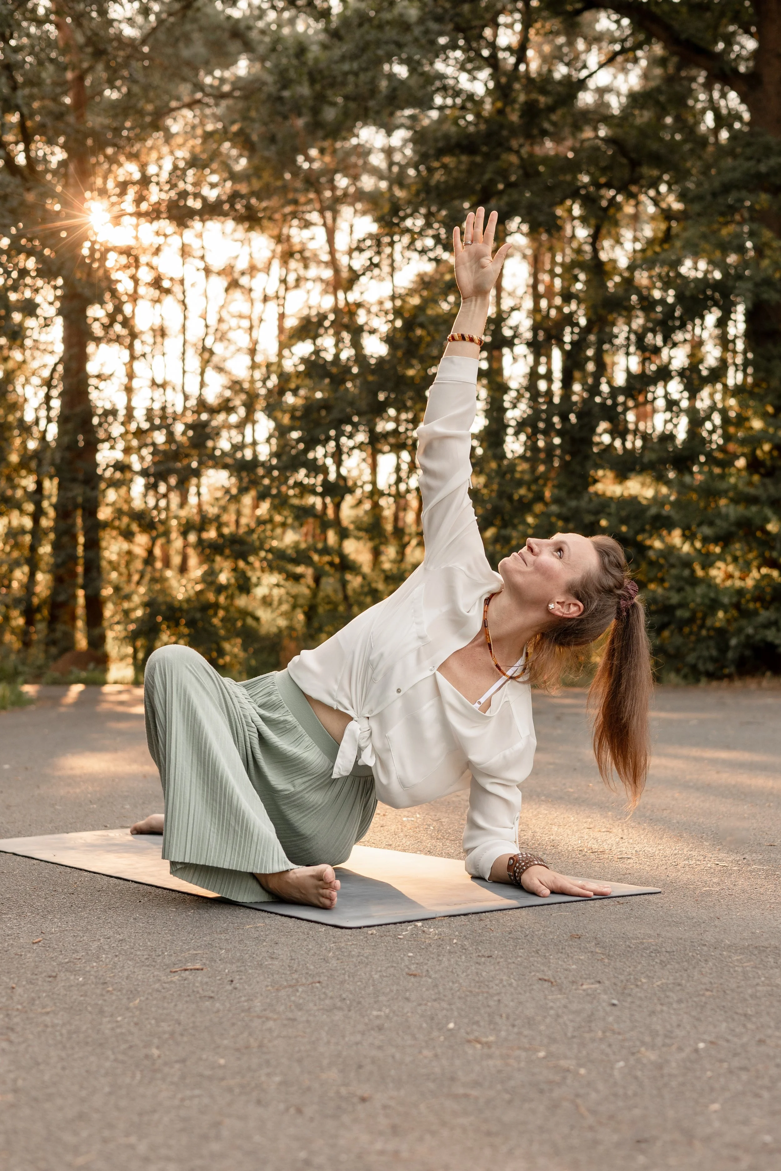 Eine Frau macht Yoga im Freien auf einer Matte, während sie auf dem Boden sitzt und in eine Richtung blickt, bei Sonnenuntergang im Hintergrund, umgeben von Bäumen.