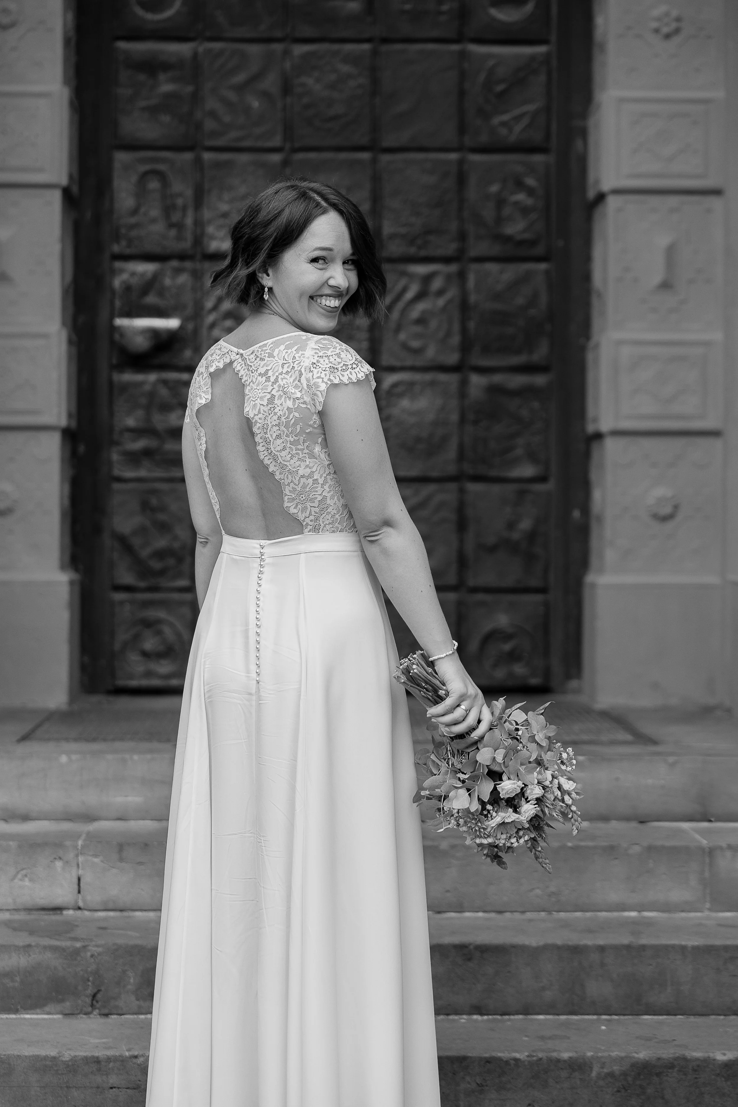 Schwarz-weiß-Foto einer lachenden Frau in einem Hochzeitskleid, die auf den Stufen vor einem großen Holztor steht, in der Hand hält sie einen Blumenstrauß.
