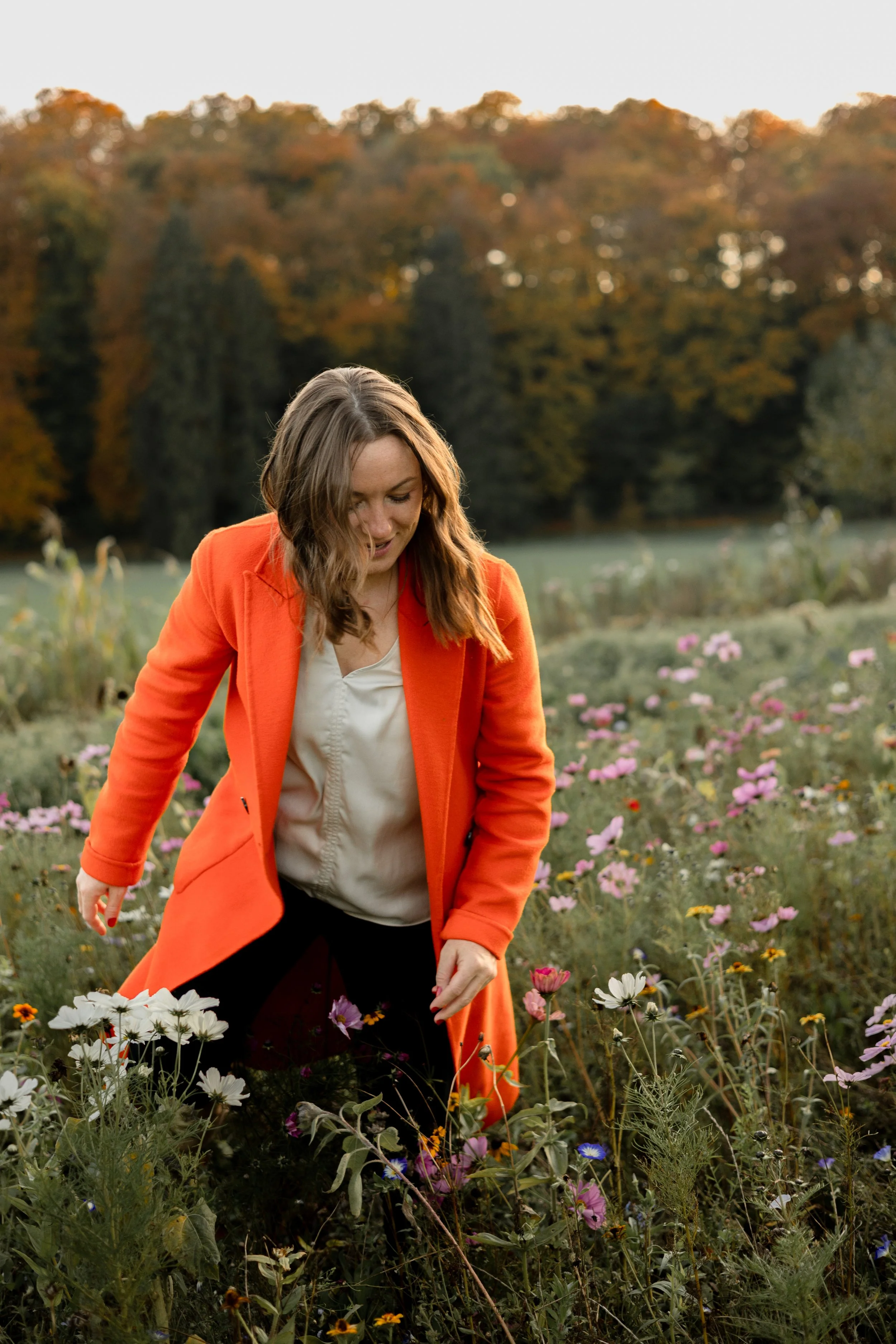 Junge Frau in orange Mantel beim Blumenpflücken in einem Garten mit bunten Blumen, im Hintergrund Bäume im Herbst.