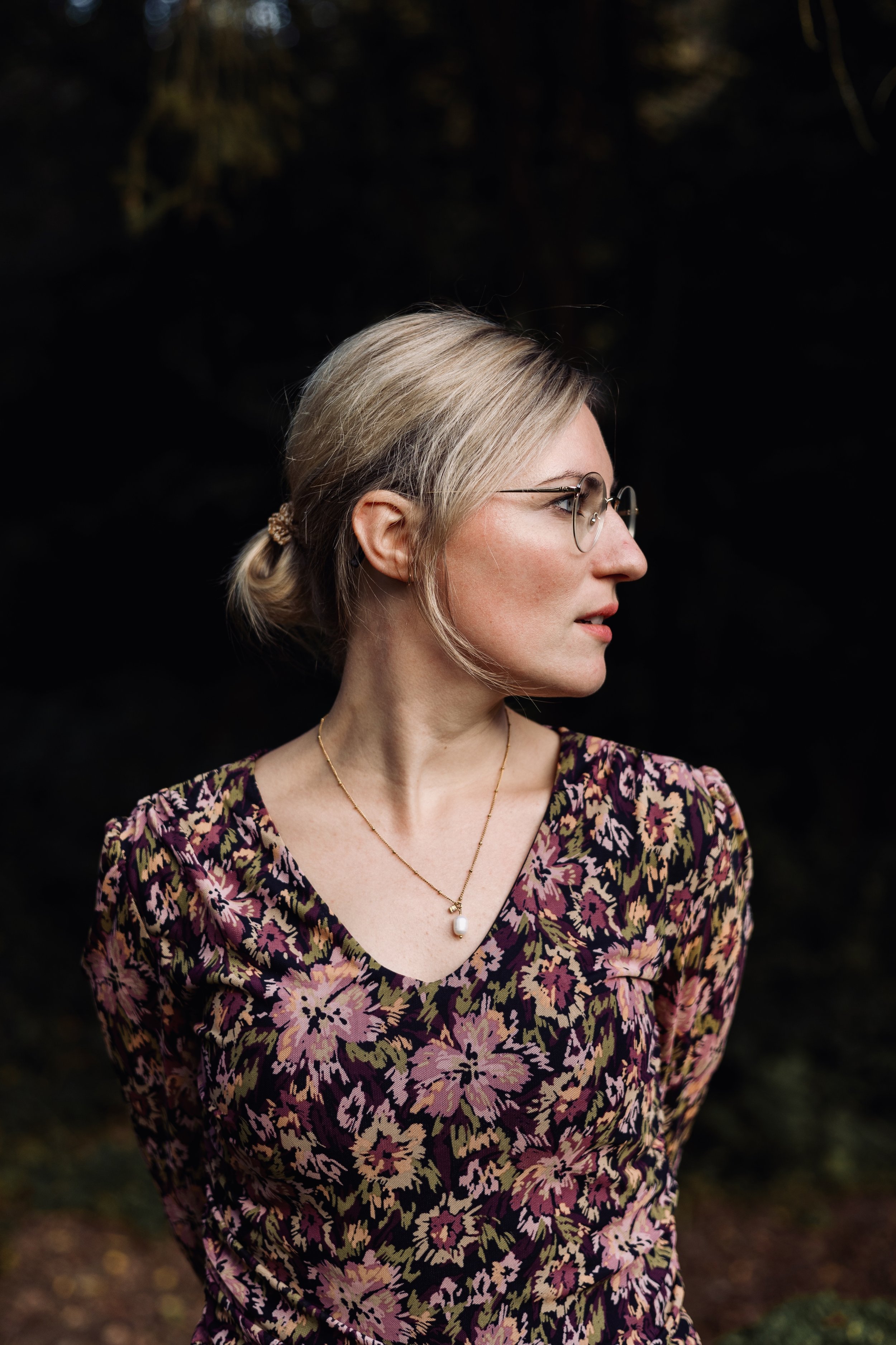 Eine Frau mit blondem Haar, die auf der rechten Seite des Bildes im Profil zu sehen ist, trägt runde Brillen, einen Blumen-Dress in dunklen Tönen und eine Perlenkette. Der Hintergrund ist dunkel und unscharf.