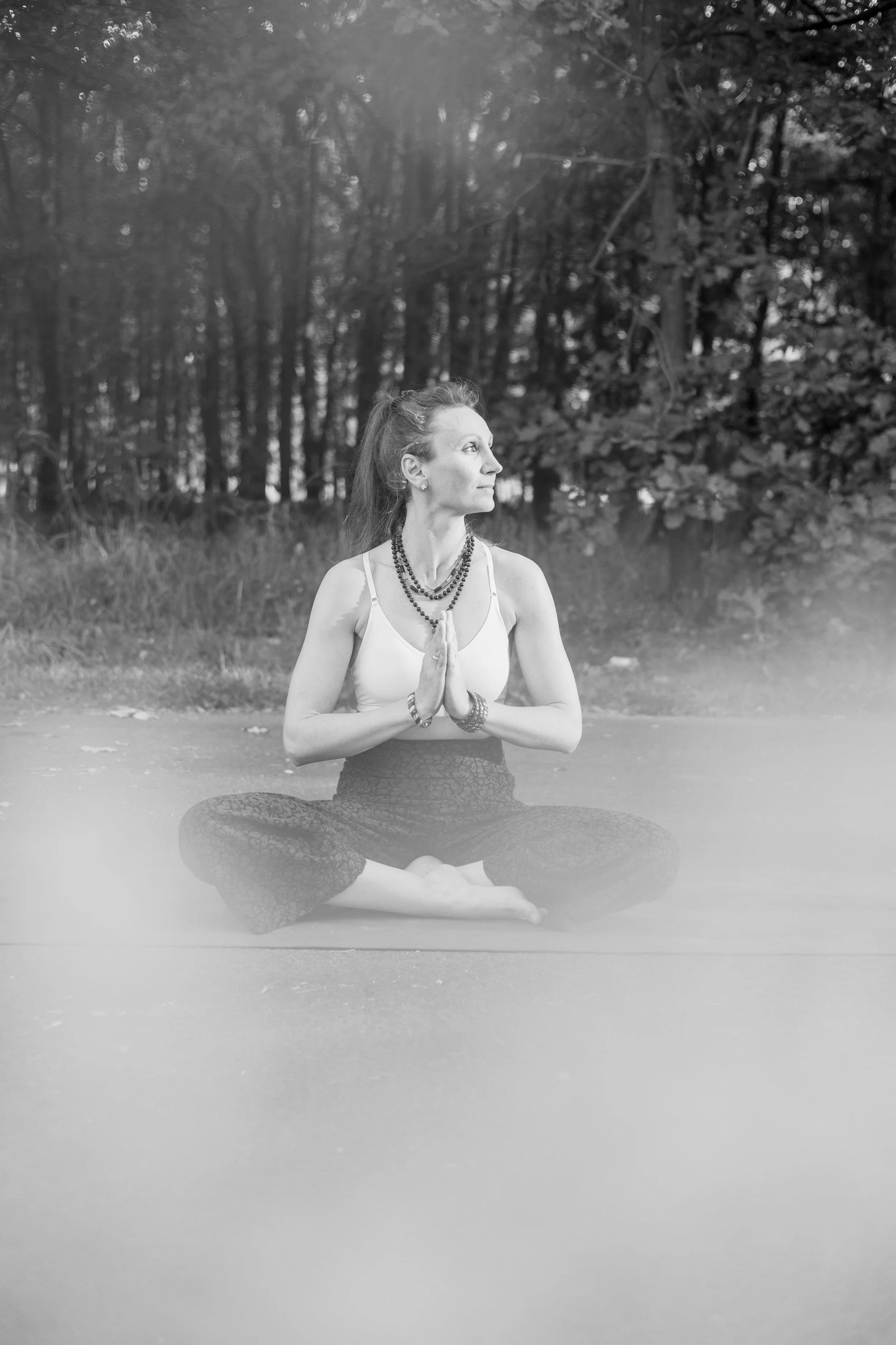 Schwarz-Weiß-Foto einer Meditierenden Frau in einer Yogapose im Freien, sitzend im Schneidersitz, mit gebeteten Händen vor der Brust, vor einem Waldhintergrund.
