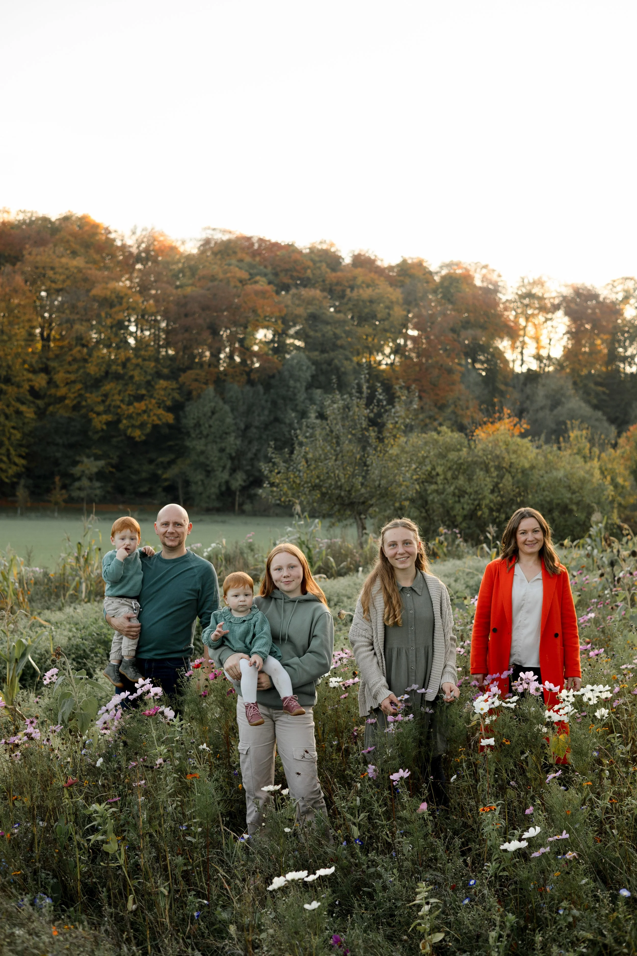 Eine Gruppe von sechs Menschen, zwei Erwachsene und vier Kinder, steht inmitten von bunten Blumen in einem Garten bei Sonnenuntergang, im Hintergrund ein Wald mit herbstlichen Bäumen.