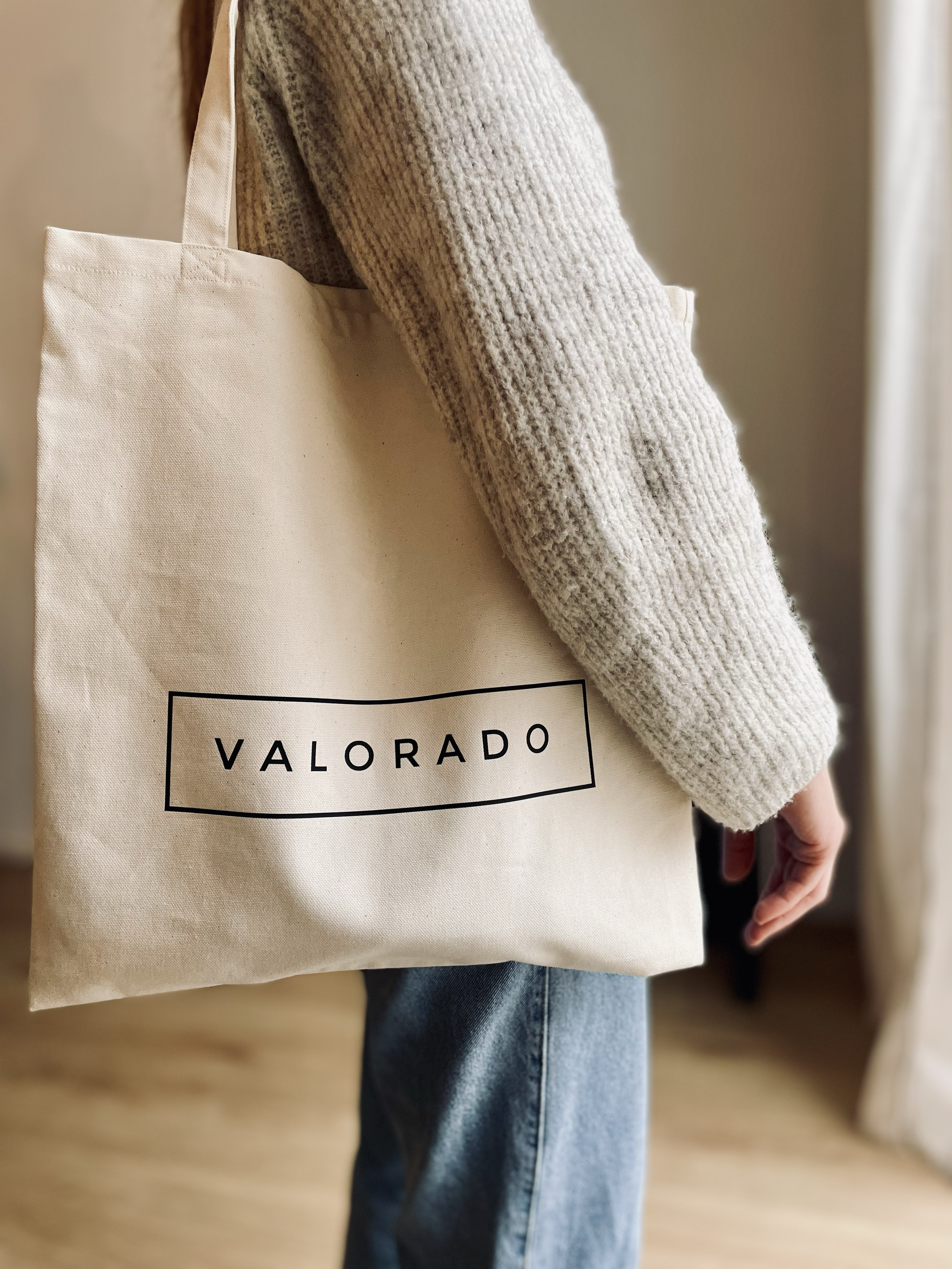 Person trägt eine beige Stofftasche mit dem Aufdruck 'VALORADO', darüber trägt die Person einen beigefarbenen Pullover und blaue Jeans.