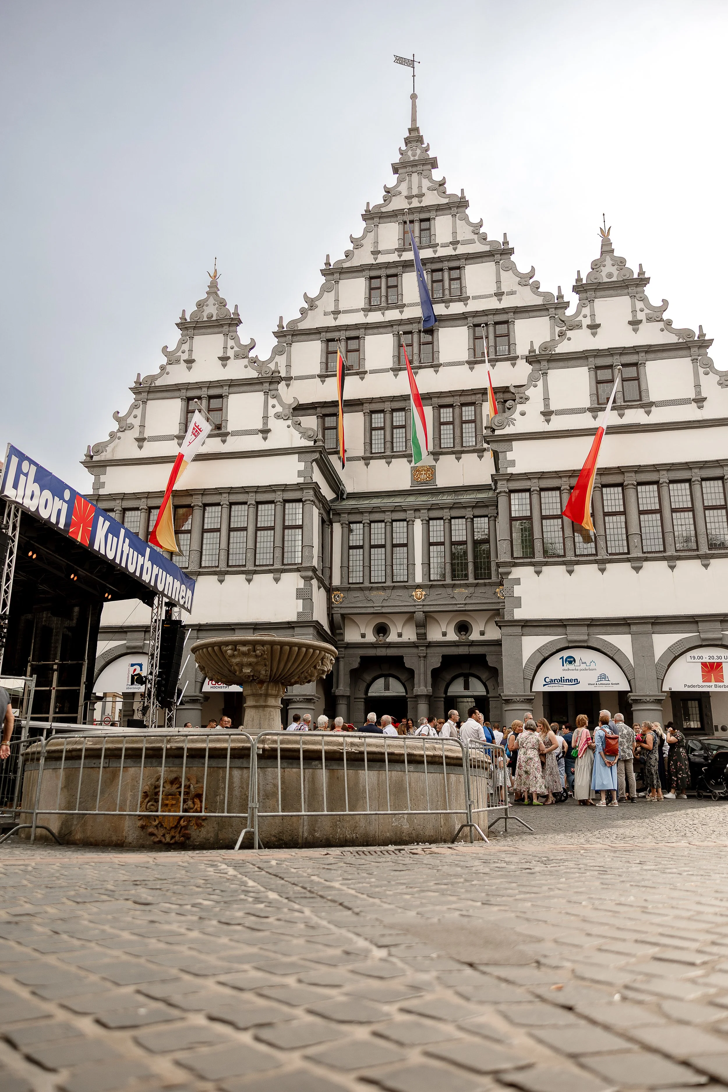 Ein historisches Gebäude im Barockstil mit mehreren Flaggen vor einer Menschenmenge, die sich in einem öffentlichen Platz versammelt hat, vor einem Brunnen und einem Banner mit dem Text "Kultur", in Deutschland.