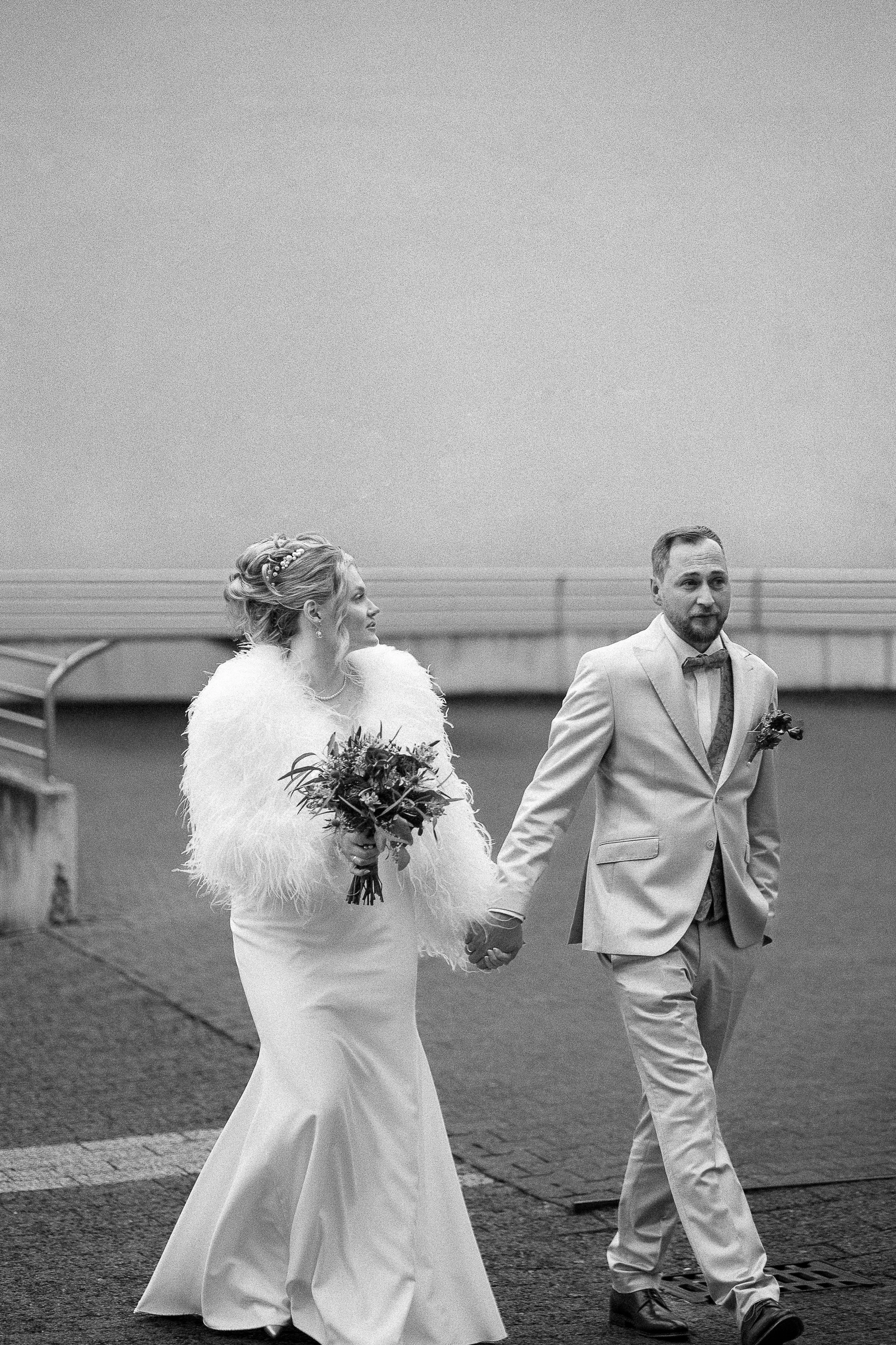 Braut in einem langen weißen Kleid mit Federjacke, Brautstrauß in der Hand, neben einem Bräutigam in einem hellgrauen Anzug und Fliege, beide Hand in Hand, gehen draußen auf der Straße.