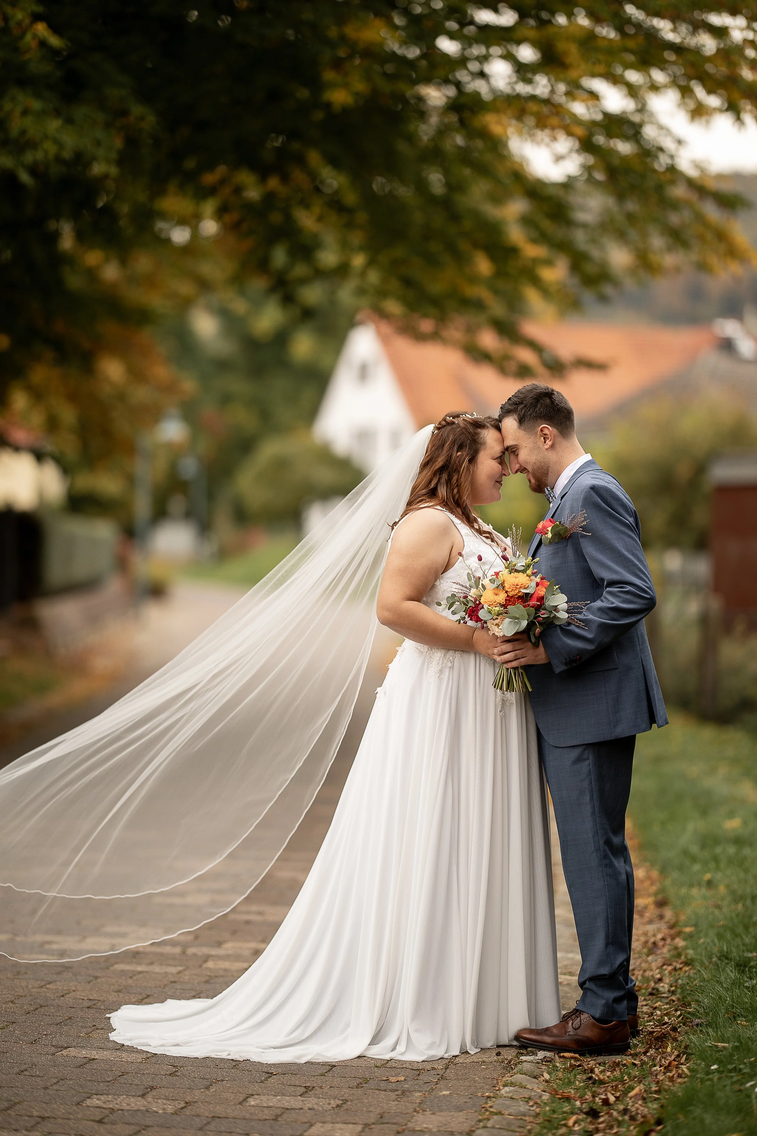 Ein Brautpaar steht verliebt vor einem Baum im Herbst, mit gestreckter Stirn und Stirn an Stirn. Die Braut trägt ein weißes Hochzeitskleid mit langem, fließendem Schleier, und hält einen bunten Blumenstrauß. Der Bräutigam trägt einen blauen Anzug, we