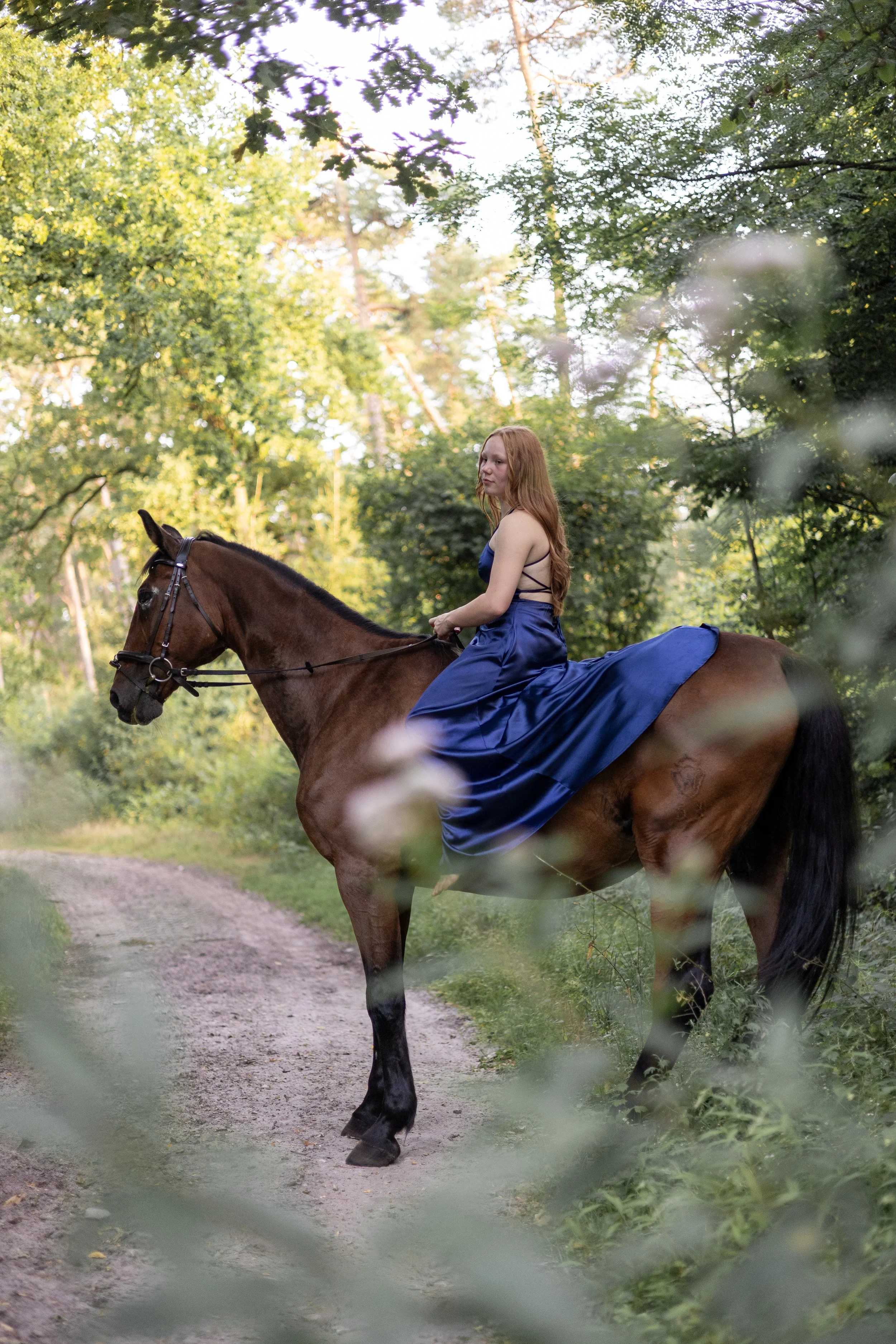 Frau mit langen roten Haaren, in einem blaue Abendrobe, reitet auf einem braunen Pferd durch einen Waldweg, umgeben von grünen Bäumen.