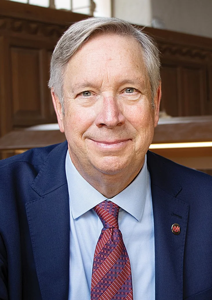 A middle-aged man in a dark blue suit, light blue shirt, and patterned red tie, smiling in a professional setting with wooden paneling in the background.