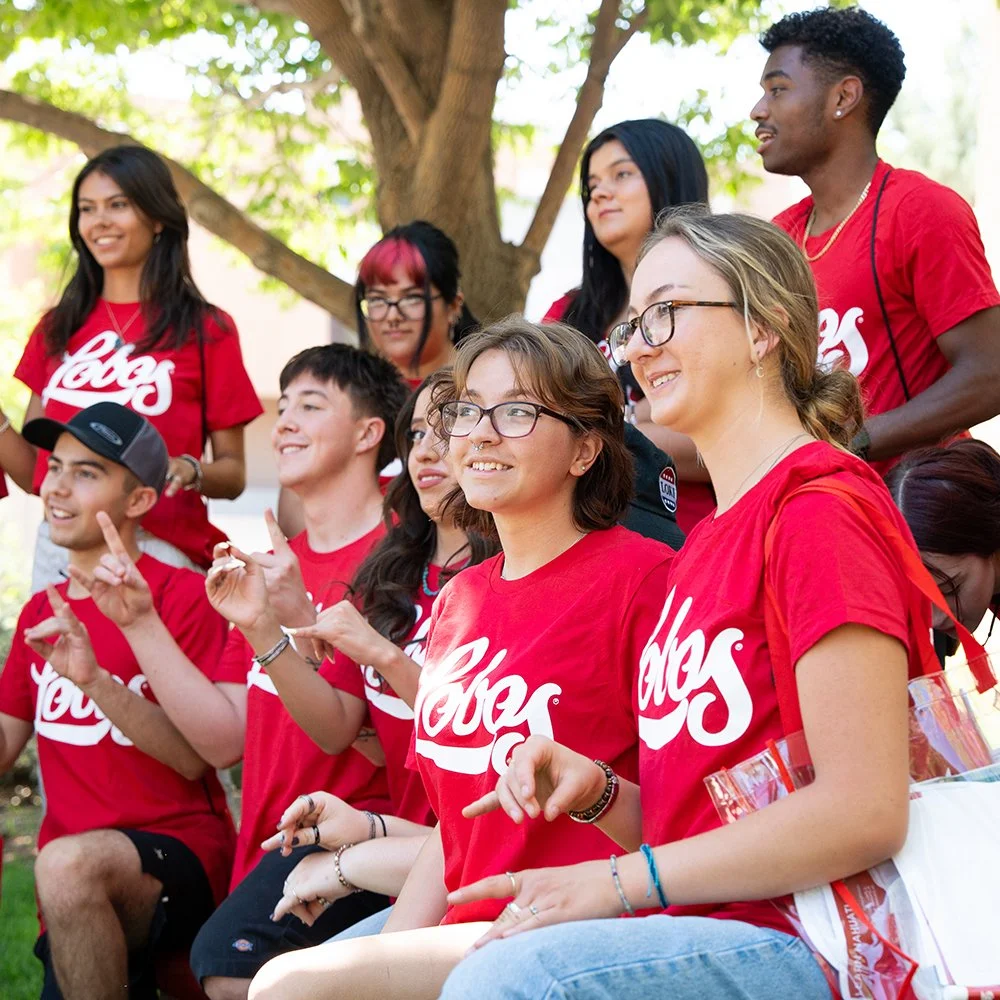 A group of young people wearing red t-shirts with the Coca-Cola logo, gathered outdoors in a park-like setting, smiling and making gestures.
