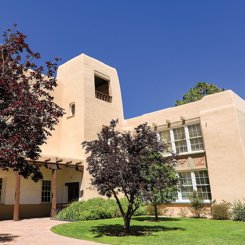 A beige adobe-style building with several windows, a small tower, and a balcony under a clear blue sky, surrounded by green trees and grass.