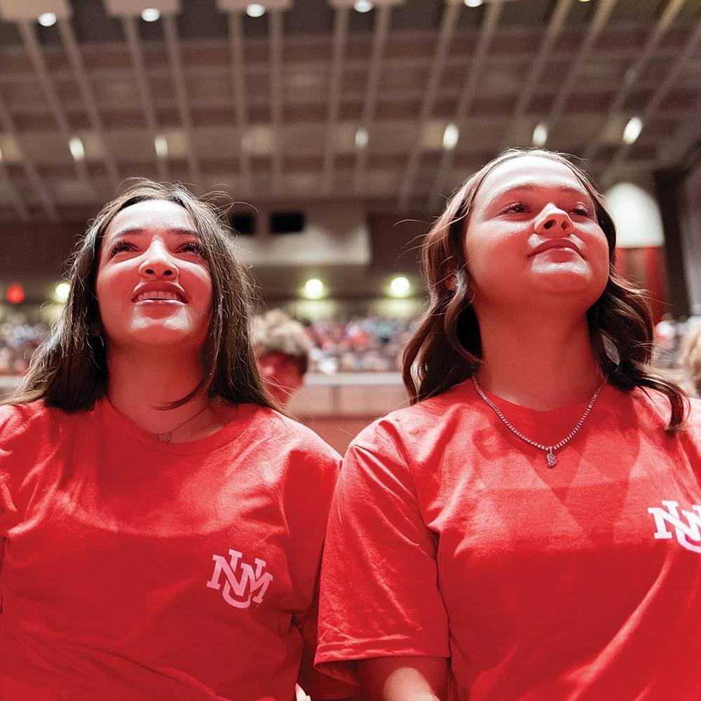 Two young women wearing red T-shirts with a white 'NY' logo sitting in a large auditorium or stadium, looking upwards with smiles.