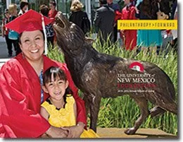 A woman in a red graduation gown and cap is smiling and sitting beside a young girl in a yellow dress. They are posing next to a bronze wolf statue. The background includes a banner reading 'Philanthropy Forward' and the 'University of New Mexico' logo.