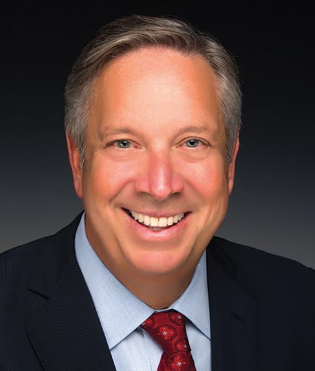 Headshot of a smiling man in a dark suit, light blue shirt, and red tie with a dark background.