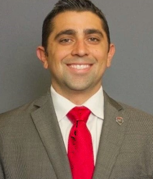 A man in a gray suit with a red tie smiling, against a plain background.