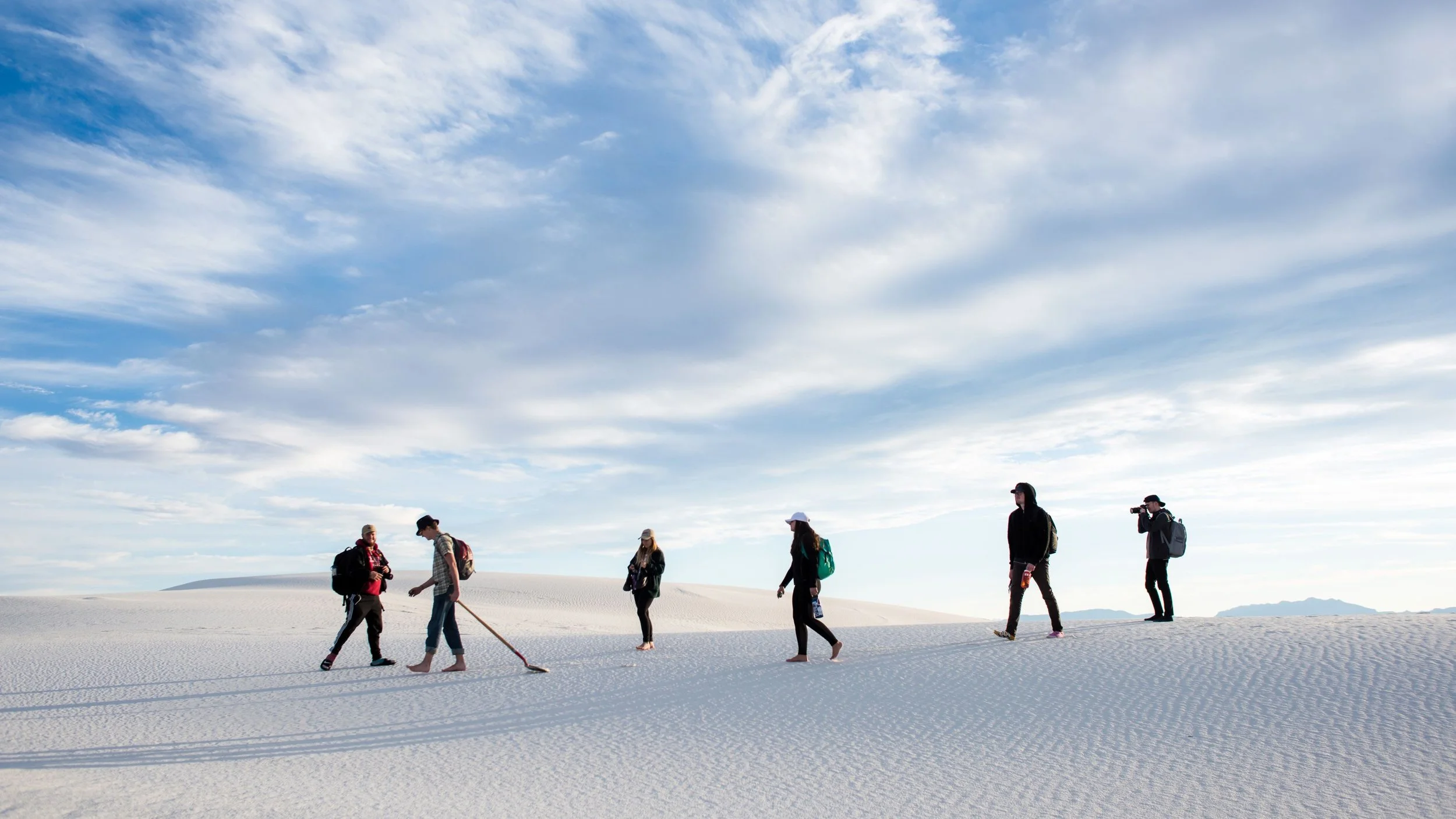 Group of seven people walking on white sand dunes under a partly cloudy sky, some carrying backpacks and one with a camera.