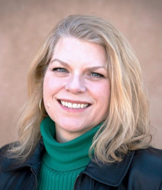 A woman with blonde hair smiling, wearing a green turtleneck and a black jacket, outdoors against a blurred background.