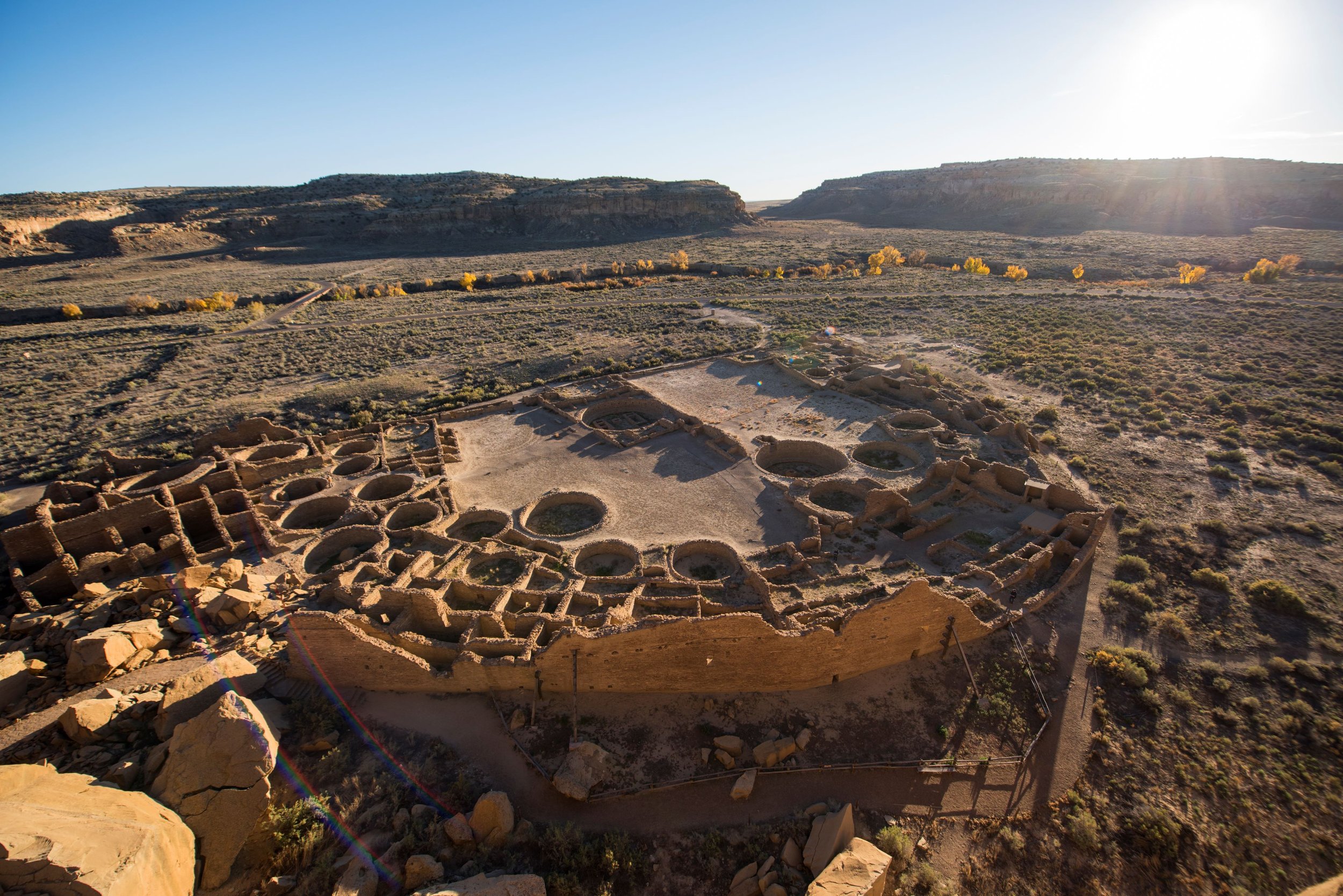 Aerial view of ancient cliff dwellings with circular and rectangular rooms, set in a desert landscape with sparse vegetation and distant mesas, under a clear blue sky.