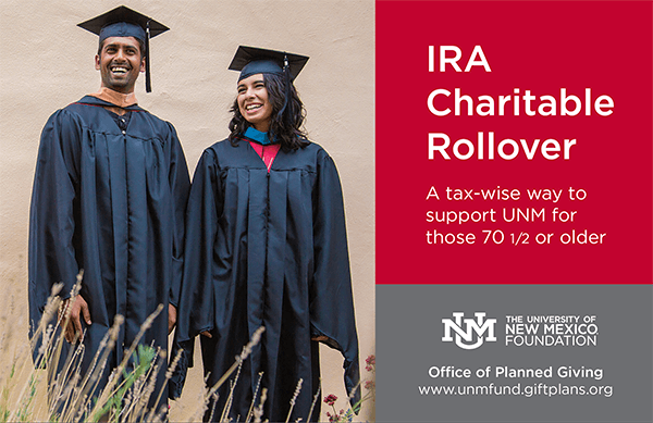 Two graduation graduates in caps and gowns standing outdoors next to a red and gray sign promoting IRA Charitable Rollover for supporting UNM, with the University of New Mexico Foundation logo.