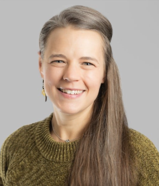 A woman with long gray hair, smiling, wearing a green sweater and a small earring, against a gray background.