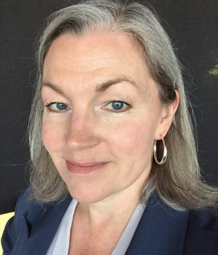 Close-up of a woman with gray hair, blue eyes, wearing hoop earrings and a navy blazer, smiling slightly.