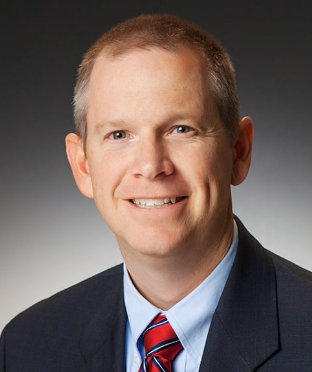 Headshot of a smiling man in a suit and tie against a dark background.