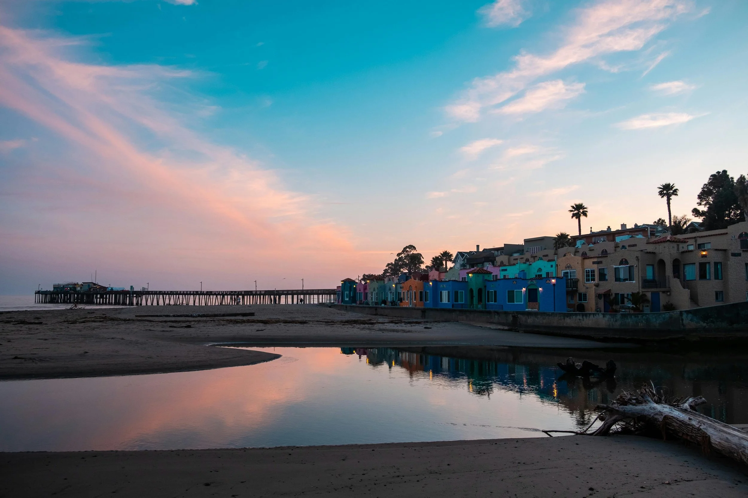 Colorful houses on a hillside along the beach with a pier extending into the ocean, reflected in a calm water pool at sunset.