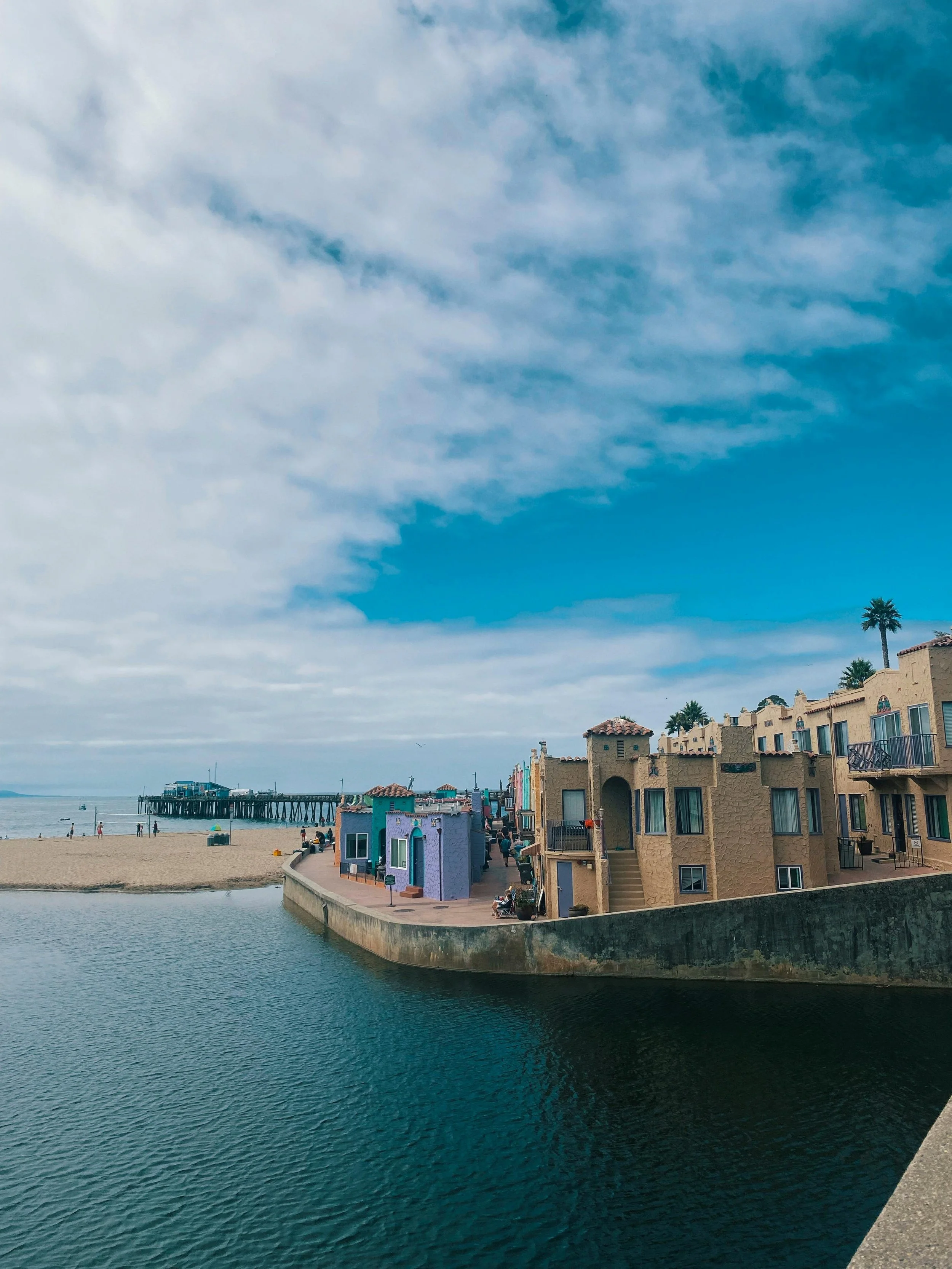 A coastal scene with colorful buildings along a waterway, a sandy beach, and a pier extending into the ocean under a partly cloudy sky.