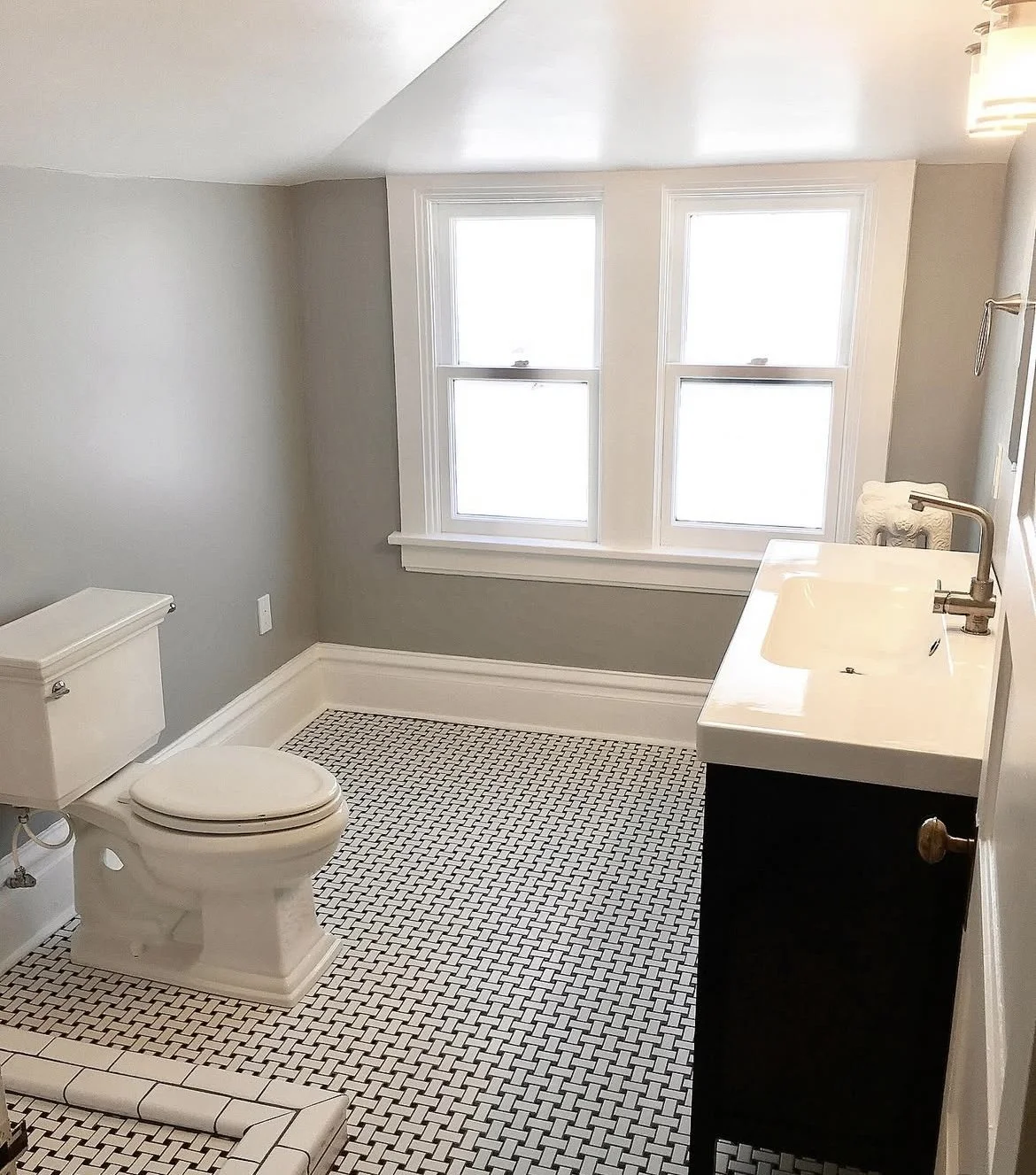 A clean, minimalist bathroom with a white toilet, black cabinetry with a white sink, and two large windows with white framing. The floor has small black and white mosaic tiles, General Contractors based out of Parker, Colorado