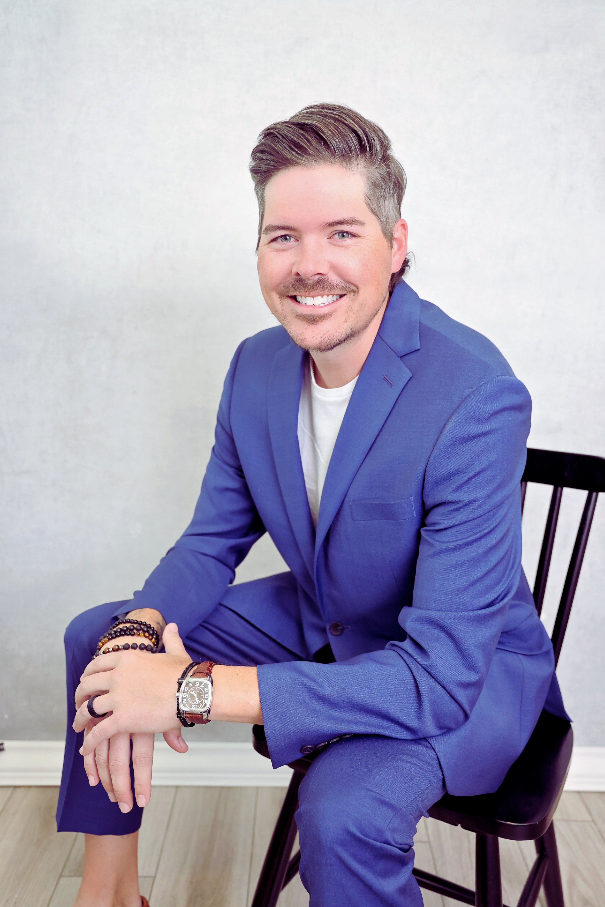A smiling man in a blue suit sitting on a black chair against a light-colored wall.