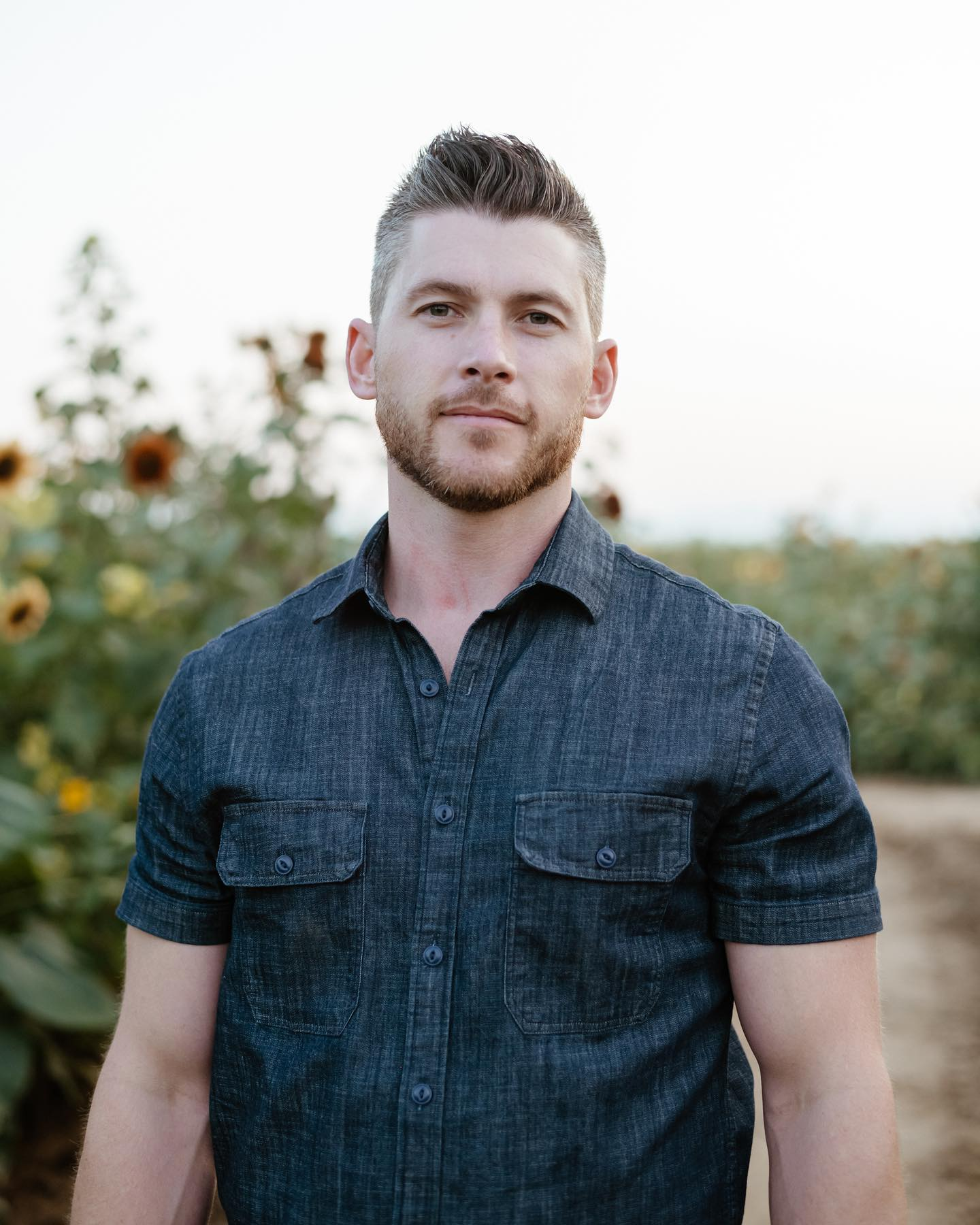 A man with short brown hair, a beard, wearing a dark denim shirt, standing outdoors with sunflower plants in the background.